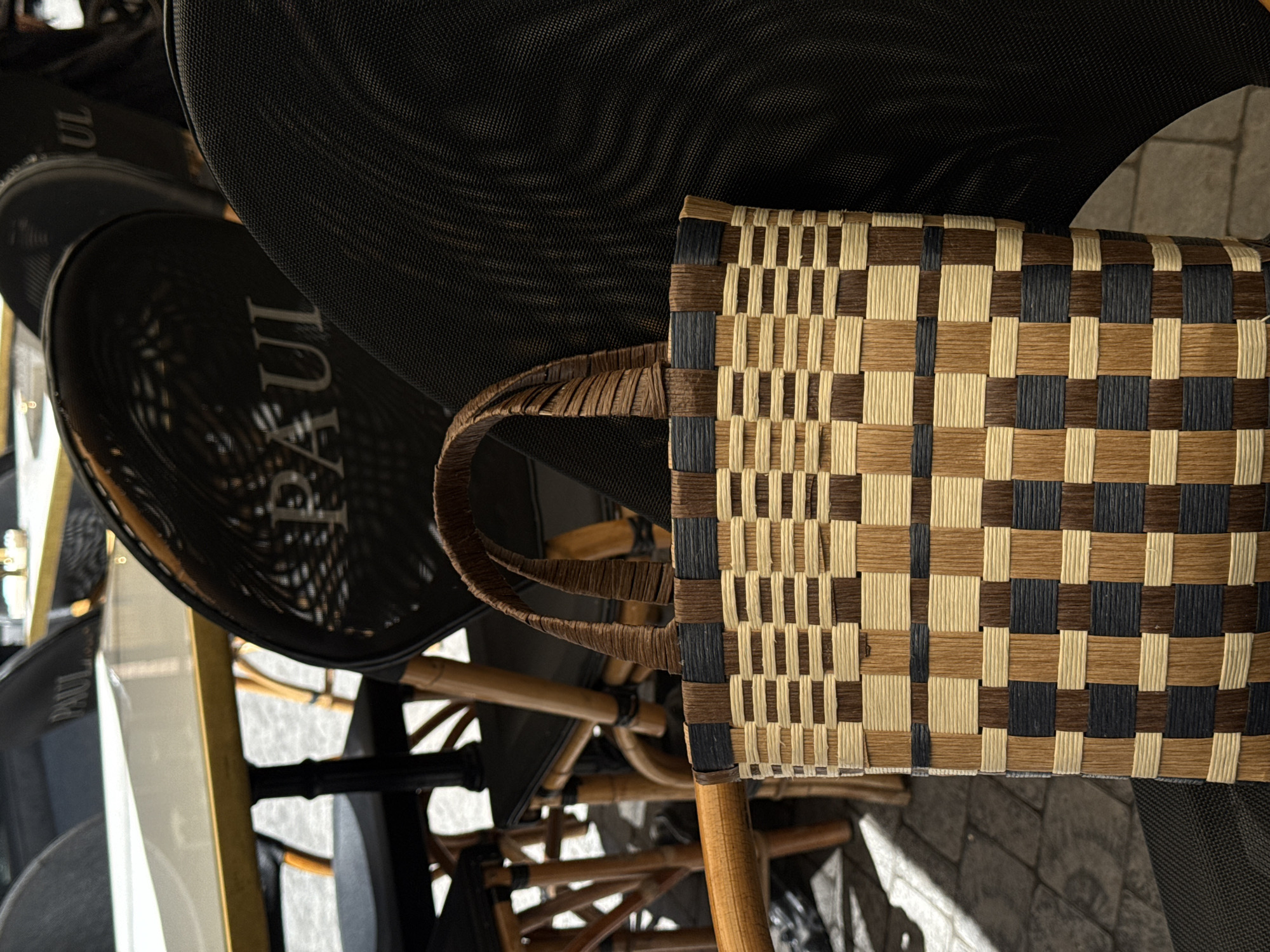 An outdoor cafe setting in Yerevan, Armenia features a woven basket bag with a checkered pattern of beige, brown, and dark blue strips. The bag rests on a black chair with a meshed back, positioned on cobblestone ground. In the background, there are tables and chairs, some with the "PAUL" logo visible on their backs. The scene is captured in daylight with natural shadows.