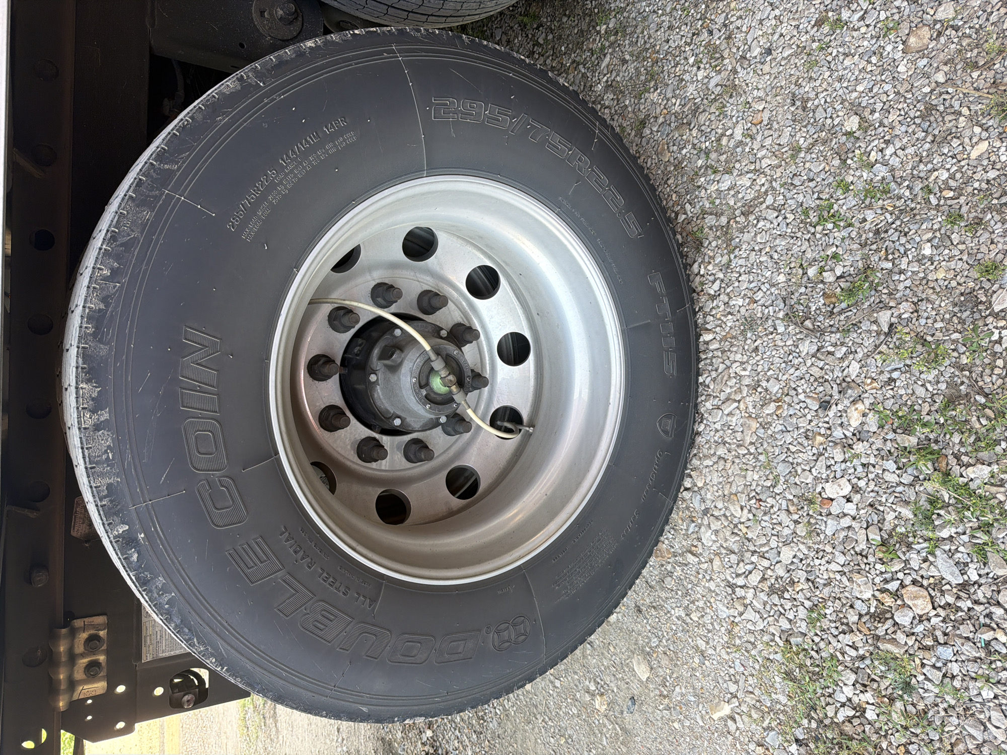 This is a close-up, eye-level, exterior, medium shot of a large truck tire mounted on a steel rim. The tire is black and shows prominent lettering which reads "DOUBLE COIN ALL STEEL RADIAL" on the left side and "295/75R22.5 144/141L 14PR" on the right side. The rim is silver and has multiple lug nuts. A white wire is attached to a green valve stem on the wheel. The background consists of gravel and some patches of green grass. The truck's undercarriage is visible in the upper portion of the image. There are no people or animals present. The location is not specified.