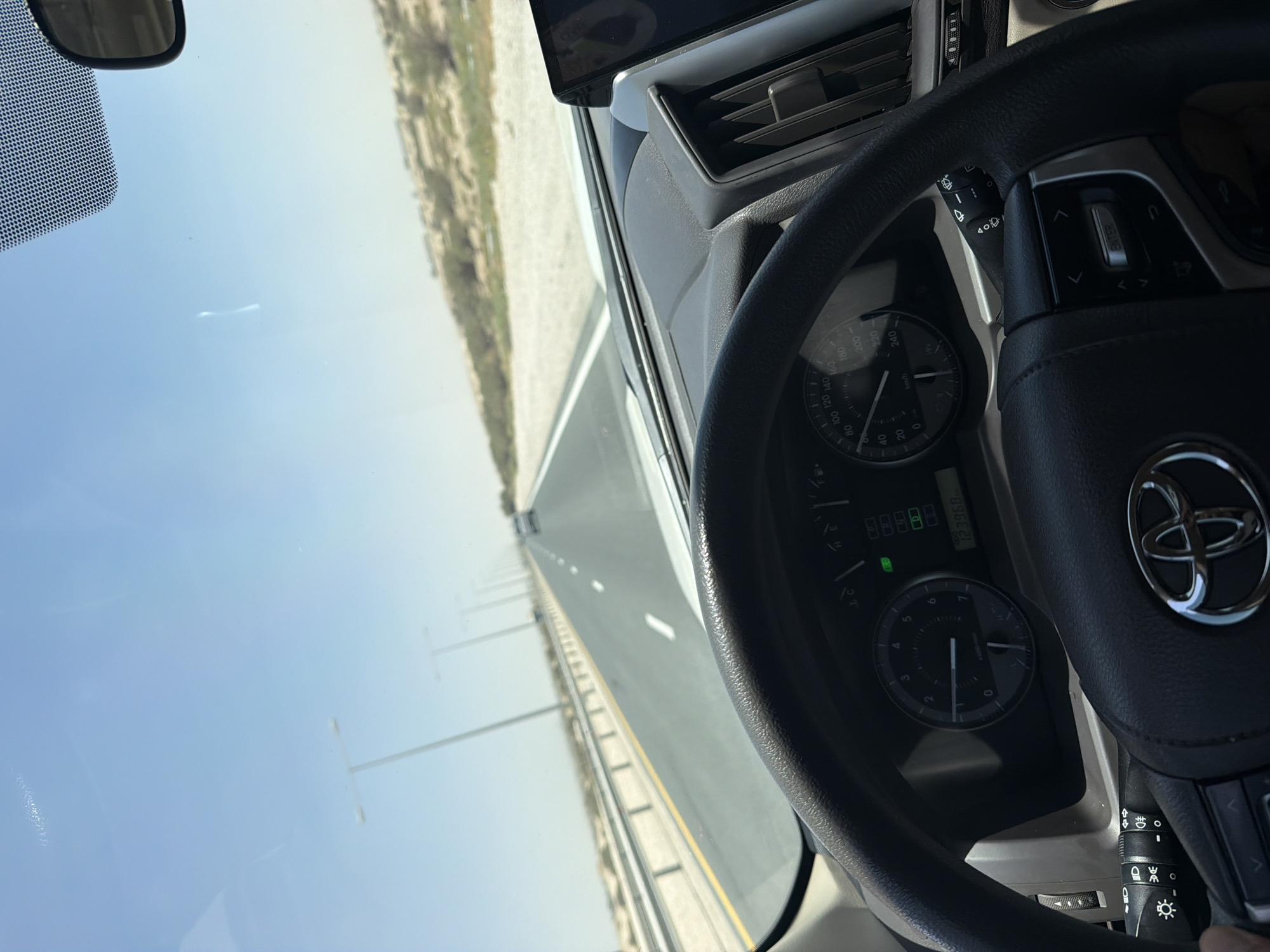 The image captures the perspective of a driver looking forward through a car's windshield. The steering wheel is prominently in the foreground, with part of the dashboard visible. The dashboard includes a speedometer, tachometer, and a digital display showing "123968" and "D". A car's rearview mirror is visible at the top right. The car is on a paved road with lane markings, bordered by a barrier and sandy terrain with sparse vegetation. In the distance, a few other vehicles and tall light poles line the road, suggesting a highway in a desert environment. The sky is clear and blue.