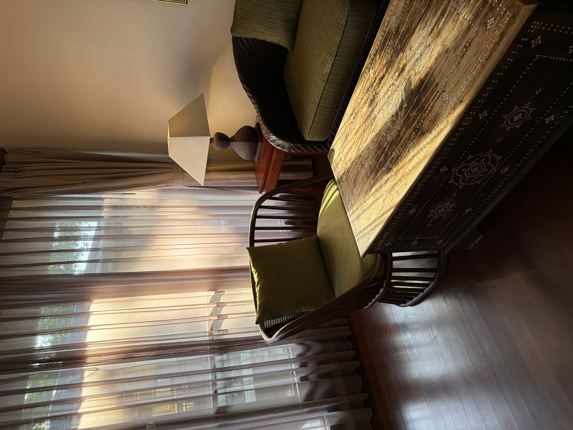 The image captures an interior room, likely a living or lounge area, imbued with warm, golden sunlight. The scene is viewed from a slightly elevated perspective, looking into the room.
In the foreground, a long, dark wooden table or bench with intricate white inlay patterns extends across the right side of the frame. These patterns consist of geometric shapes, starbursts, and circular motifs, illuminated brightly by direct sunlight, highlighting their detailed craftsmanship.
To the left of this ornate table, a wicker armchair with a green seat cushion and matching throw pillow is positioned, facing towards a large window. Further back, another piece of furniture, a sofa or larger armchair made of a dark woven material with green cushions, is partially visible.
A prominent window occupies the left side of the room, covered by sheer brown or tan curtains that filter the incoming sunlight. The light streams through these curtains, creating distinct horizontal patterns of illumination and shadow across the dark wooden floor and various surfaces. Through the translucent curtains, hints of green foliage from outside are discernible.
A table lamp with a distinctive geometric, possibly pyramid-shaped, lampshade rests on a small wooden side table situated between the window and the darker woven sofa. The lamp is unlit. The walls of the room are a plain, light color, providing a soft backdrop to the darker furniture and floor.
The absence of people suggests a quiet and tranquil atmosphere. The strong, low-angle sunlight indicates either early morning or late afternoon, contributing to a serene and inviting mood. There is no visible text in the image. This interior setting aligns with a relaxing environment found in a location like Kuta, Indonesia.