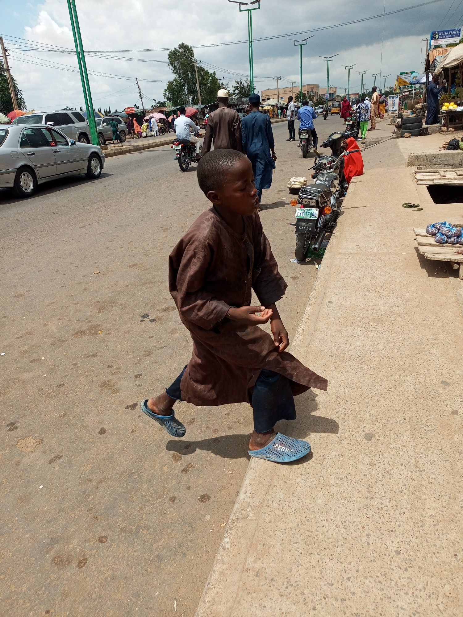 In Tempure, Nigeria, a young boy wearing a brown, patterned garment and dark pants, walks on the side of a paved road. He has on blue, open-back sandals with the word "ZEBRA" embossed on them. The boy is looking to his right, with his right hand slightly raised. The scene is outdoors on a sunny day with a cloudy sky. In the background, there are various vehicles, including cars and motorcycles. People are seen walking and some are engaged in commerce at roadside stalls. The environment appears to be a busy urban area, with a prominent traffic light pole with multiple lights on the left side of the image. A street sign with "SPRING MEDICAL DIAGNOSTIC CENTER" is visible on the right. There are also stacks of tires and merchandise displayed on wooden platforms.