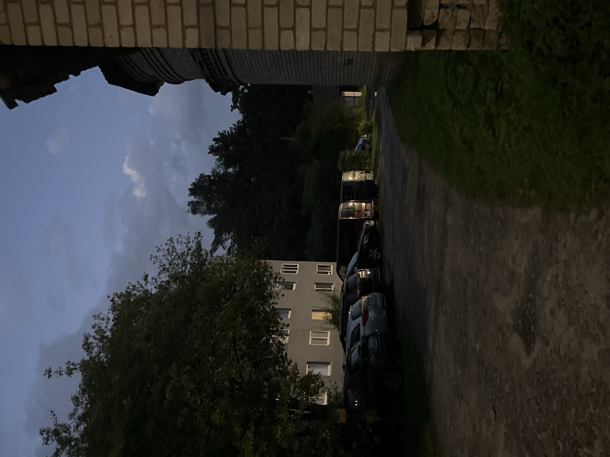 This is a night photograph taken in Vilnius, Lithuania. The image shows a parking area in front of a grey apartment building, with several cars parked. To the right of the building, a portion of a brick structure is visible, possibly part of another building or a historical edifice. The sky is dark with some clouds. The lighting suggests it is dusk or early night.