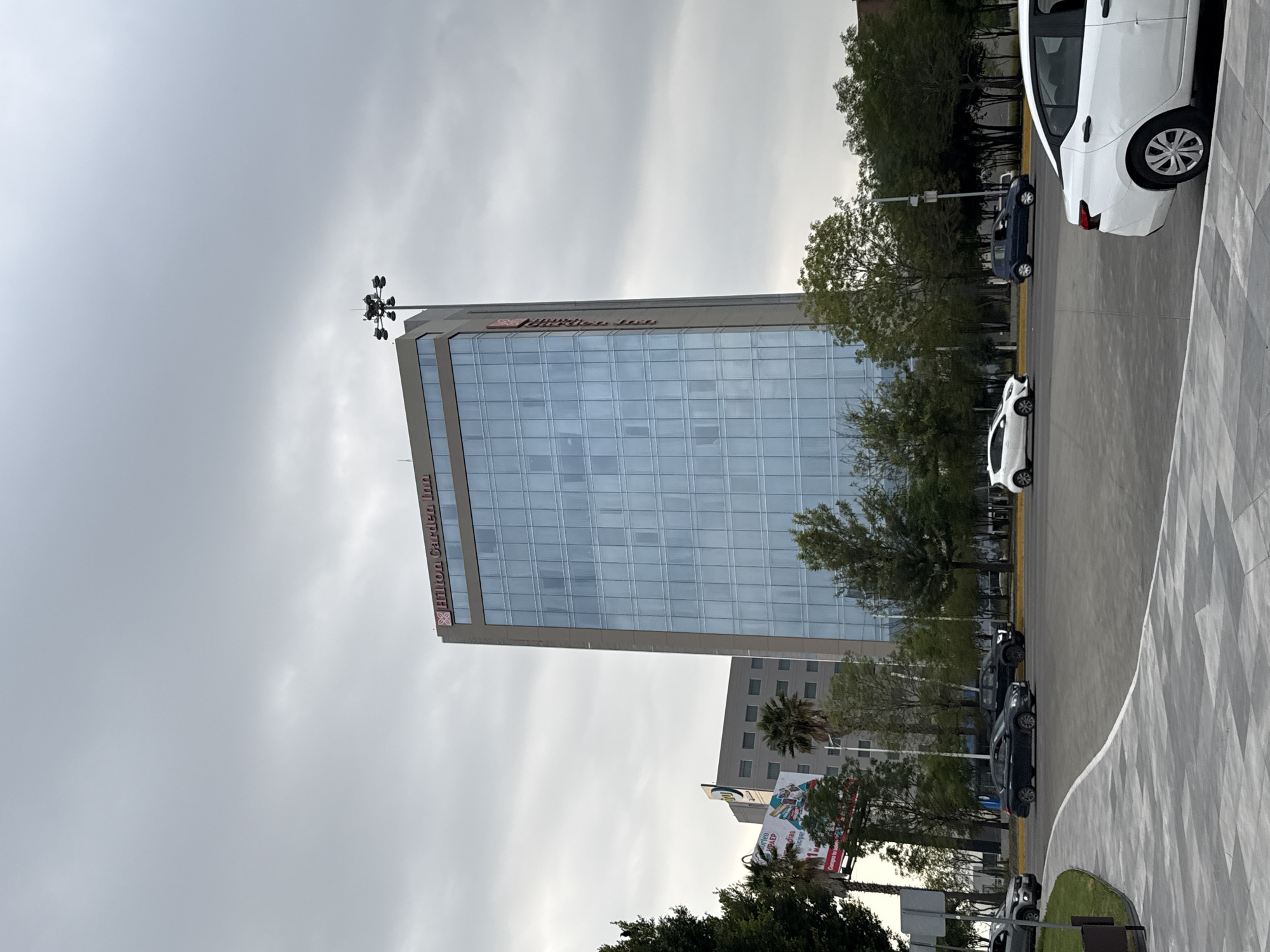 The image shows a tall, modern building in San Bernardino Tlaxcalancingo, Mexico. It appears to be a Hilton Garden Inn, identifiable by the logo on the side of the building. The building has a reflective glass facade with a grid pattern, and is surrounded by trees and some lower buildings. There are several cars parked along the side of the road near the building. The sky is overcast and gray. A light post is visible on top of the building. A large poster can be seen near the lower buildings in the area. The sidewalk has a grey and white tiled pattern.