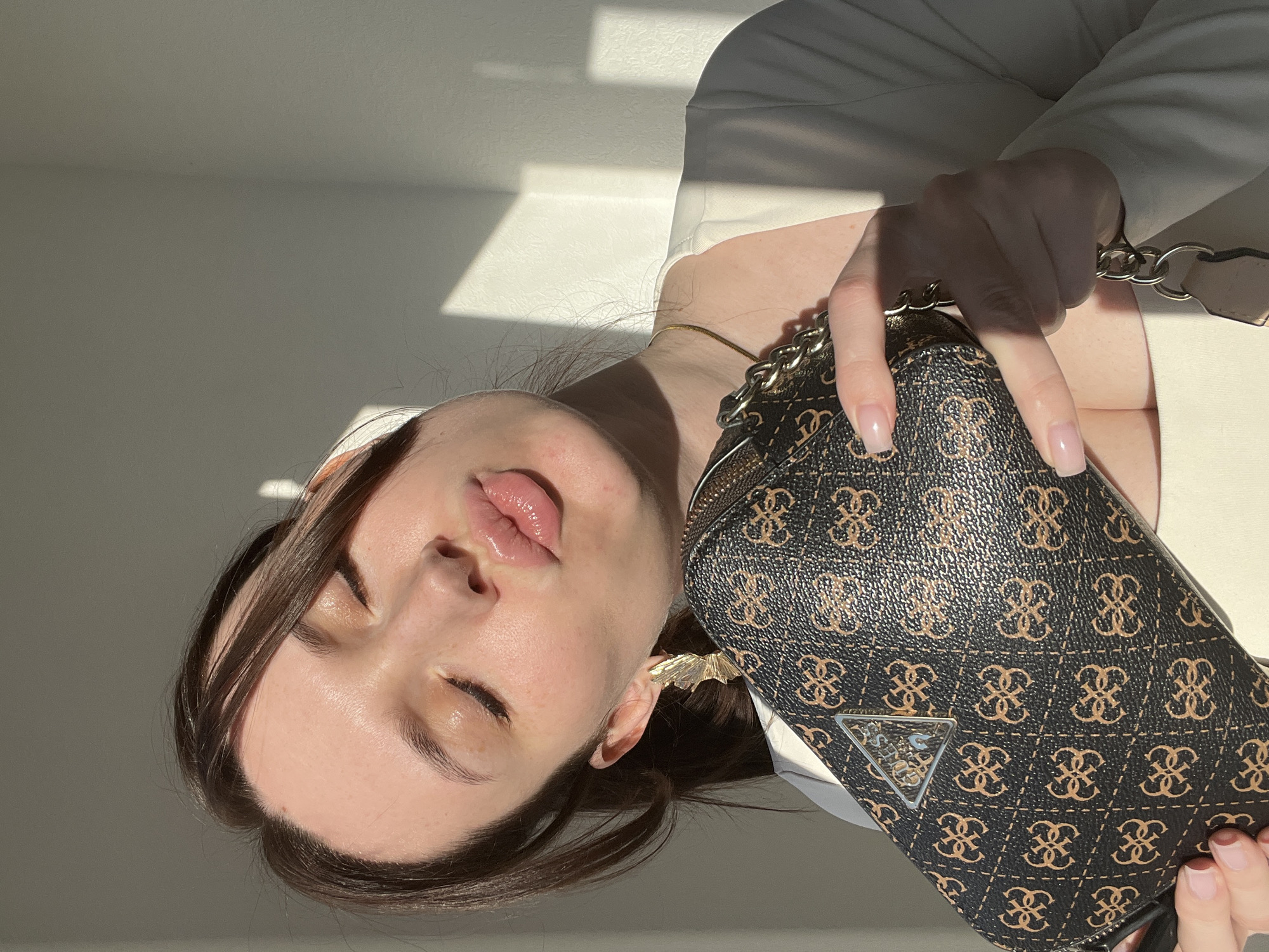 A woman with brown hair and a light complexion is holding a brown and black Guess handbag. She is wearing a white blazer. The woman's eyes are closed, and her lips are glossy. She is wearing a gold necklace and earrings. The background is a white wall with a shadow cast on it. The location is unknown, and there is no indication of Tyumen, Russia.