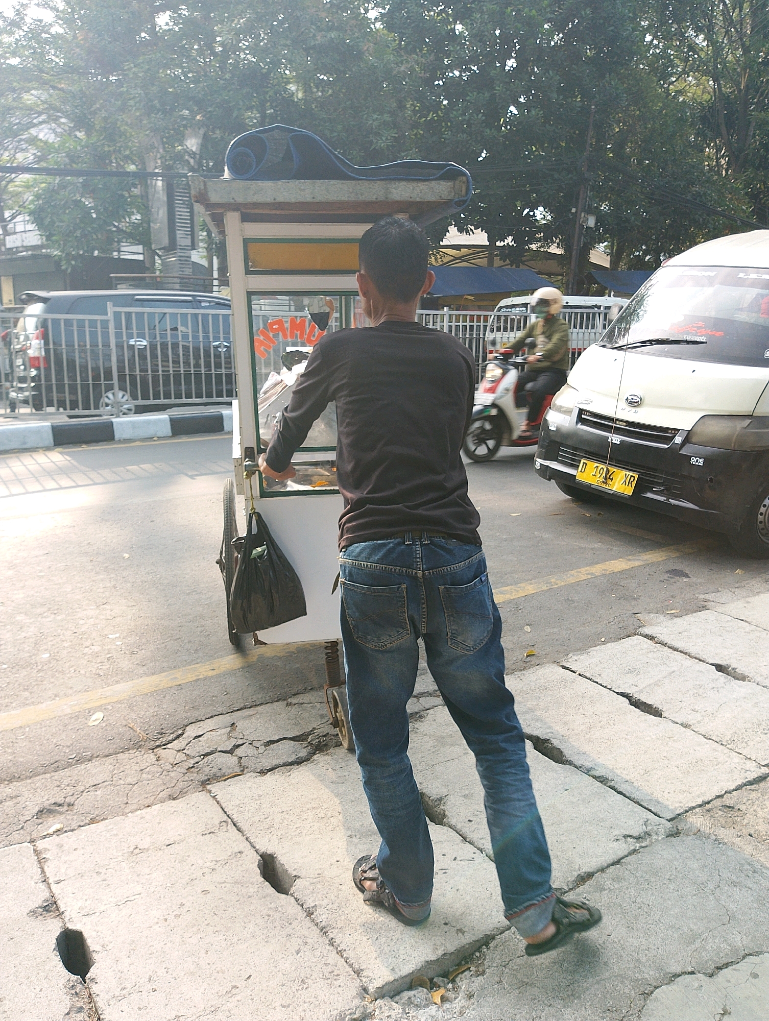 A man wearing a black long-sleeved shirt and blue jeans is pushing a food cart along a cracked concrete sidewalk in Bandung City, Indonesia. The food cart has a white exterior and is equipped with wheels. A black plastic bag is hanging from the cart. In the background, a white van with a yellow license plate that reads "D 1994 XR" is parked on the street. A person wearing a green jacket and a helmet is riding a motorcycle in the street. Trees and electrical wires are visible in the background. The scene appears to be a street-level view on a sunny day.