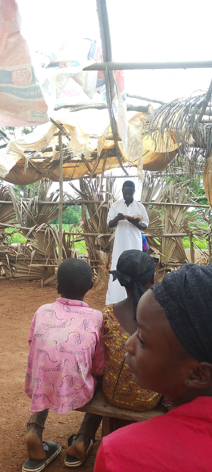 The image depicts an outdoor gathering in Kamantan, Nigeria. A man, wearing a white garment, stands in the center, holding what appears to be a book or papers. He is surrounded by several individuals, including two children and at least two women, who are seated on a wooden bench. The structure behind the man is rudimentary, constructed from branches and palm fronds, with what looks like salvaged tarpaulins or fabric serving as a roof. The ground is reddish earth. The overall setting suggests a rural or semi-rural environment, possibly a community gathering or an informal educational session.