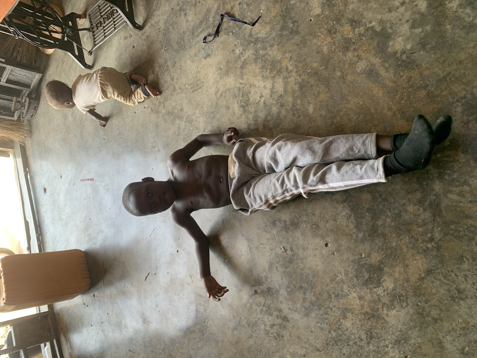 A photo taken indoors depicts two children in what appears to be a room with a concrete floor. One child is lying on their back wearing light gray pants and black socks, with their torso bare. The second child is crawling on the floor wearing a light-colored top and pants. In the upper left of the frame is a sewing machine, along with other items that could be furniture or storage containers. A blue object, perhaps a strap, is also visible on the floor. The image appears to be taken from a high angle, looking down at the children. The city of Jos, Nigeria is stated as the location.