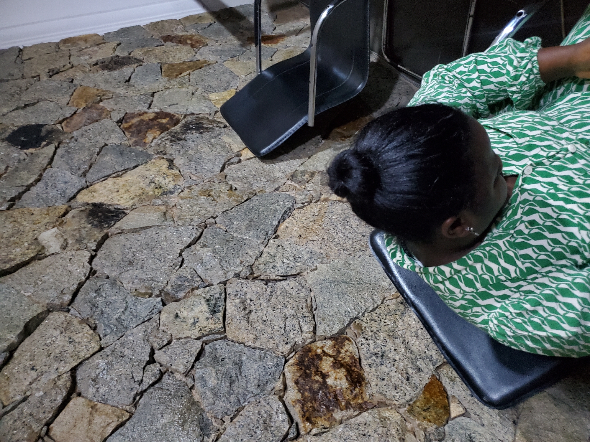 A woman with her hair pulled into a bun is seated in a black chair, facing away from the camera. She is wearing a green and white patterned dress. Behind her, a wall is covered in rough, irregular stones of varying shades of grey and brown. To her right, another black chair is visible. The scene appears to be indoors, possibly in a waiting area or lounge. The lighting is dim, casting shadows on the stone wall and the chairs.