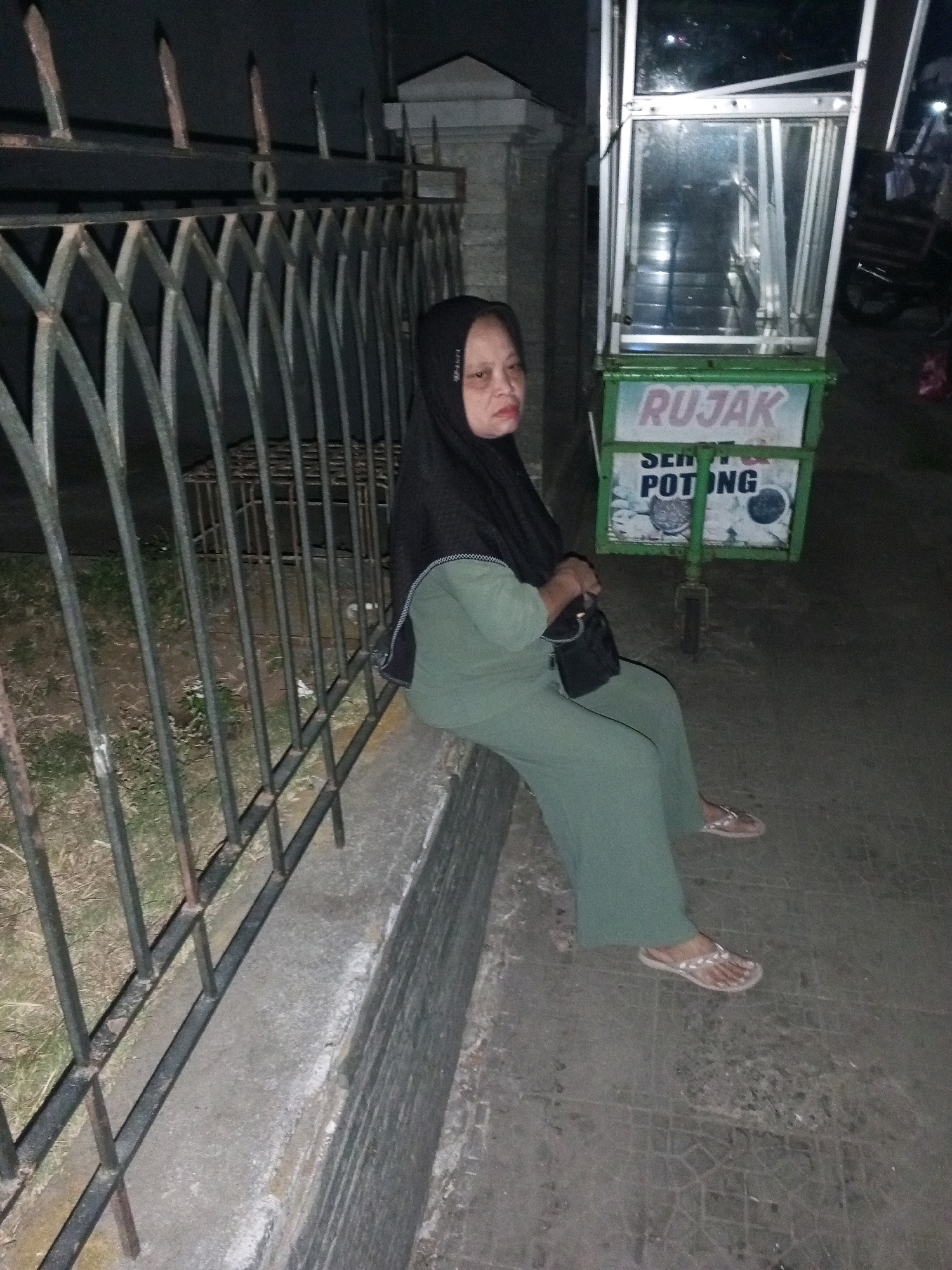 A woman wearing a hijab and a green outfit is sitting on a low concrete wall next to a black metal fence. She is holding a black handbag. To her right is a green and white food cart with "RUJAK" and "SEM" and "POTONG" written on it. The scene appears to be outdoors at night, with lighting from the food cart and possibly nearby streetlights. The ground is paved with what looks like concrete tiles. The woman is looking towards the camera.
