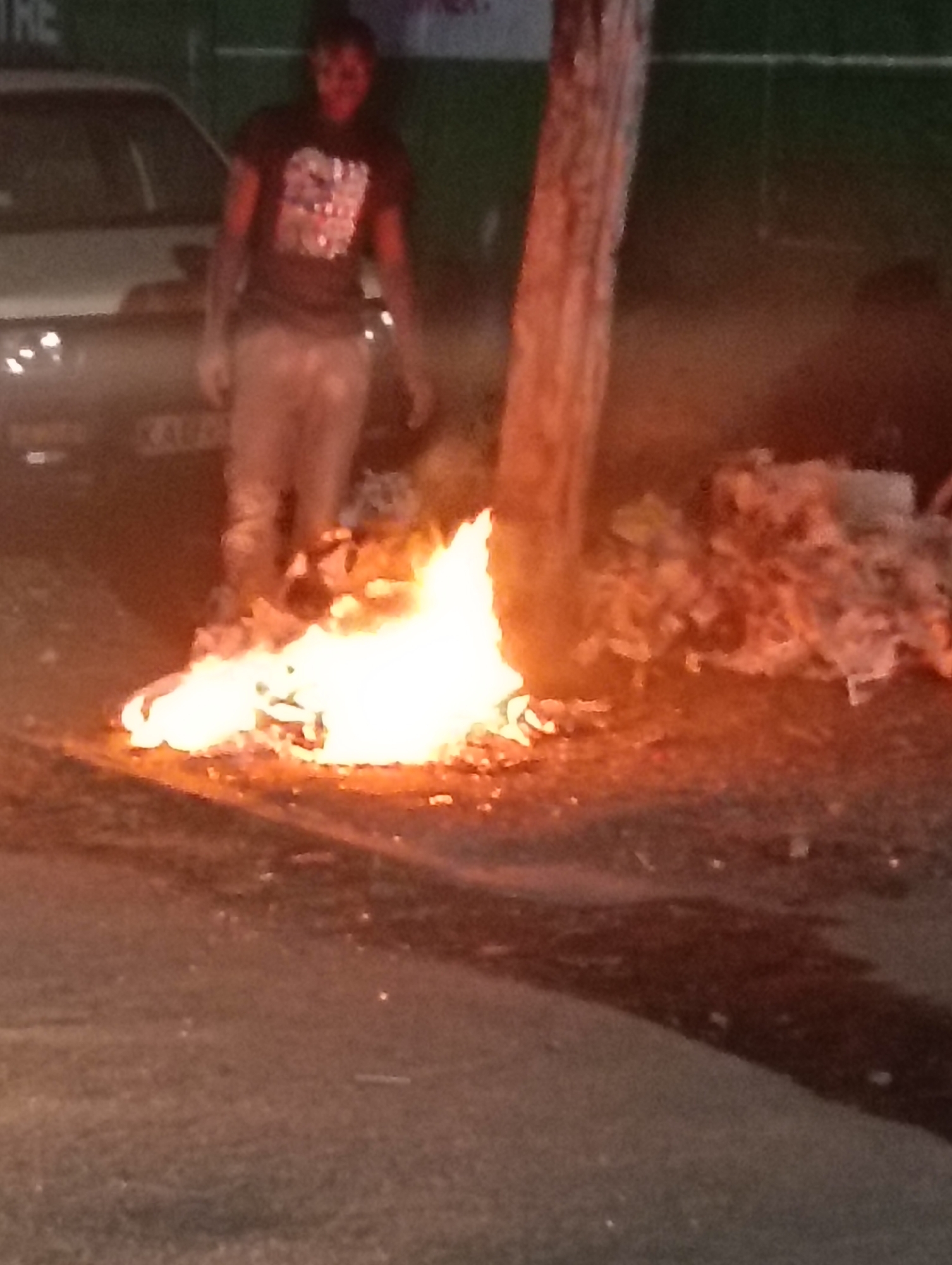 The image depicts a fire burning on a paved surface in what appears to be an urban setting. Next to the fire, there is a pile of trash and a tree trunk. A dark-skinned person wearing a t-shirt and light-colored pants stands behind the fire. A silver car is visible in the background with a license plate that reads “KAU8…” The location is plausibly Nairobi, Kenya.