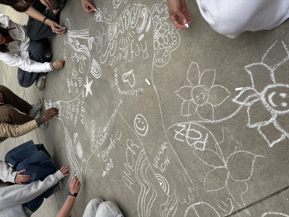 The scene depicts an outdoor activity on a gray concrete surface in the city of Unknown, Unknown. Multiple individuals are engaged in chalk art, visible from the waist down, crouched or seated on the ground around a central drawing area. At least five sets of hands are actively drawing with white chalk. The ground is extensively covered with white chalk designs, including abstract patterns, spirals, hearts, stars, and stylized flowers, some with smiling faces. Spanish words written in chalk include "AMOR" (love), "DESCANSO" (rest), "DIVERSION!" (fun!), "Hola!", "LILIA", "auto conocimiento" (self-knowledge), "Reflexion" (reflection), "Paz" (peace), and "Relación". Several small, worn pieces of white chalk are scattered on the ground, with others being held for use. Participants are dressed in casual attire, consisting of jeans, hooded sweatshirts, and sneakers. One individual wears a black wristband, and another displays silver rings on their fingers.