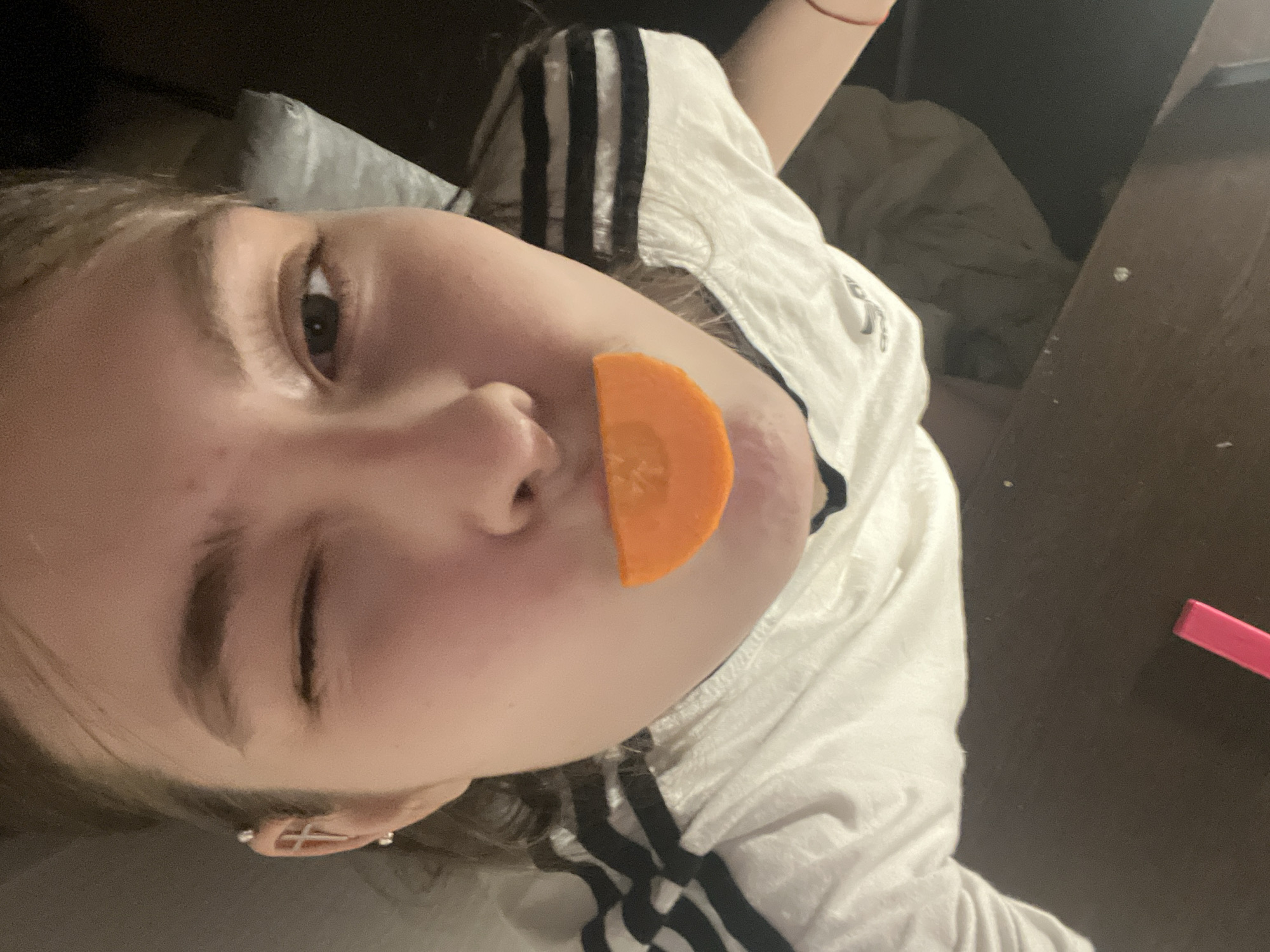 A close-up, angled selfie shows a young person with fair skin and brown hair, who is winking their left eye. A semi-circular slice of carrot is held against their upper lip, acting as a substitute for a mustache. The person is wearing a white t-shirt with three black stripes down the sleeves, likely an Adidas branded shirt. They have an earring in their left ear. The background is dark and indistinct, suggesting an indoor setting. A bed pillow is visible in the blurred background. A pink rectangular object is situated near the bottom right of the frame. Based on the visual data provided, there's no explicit indication to identify the location as Saint Petersburg, Russia.
