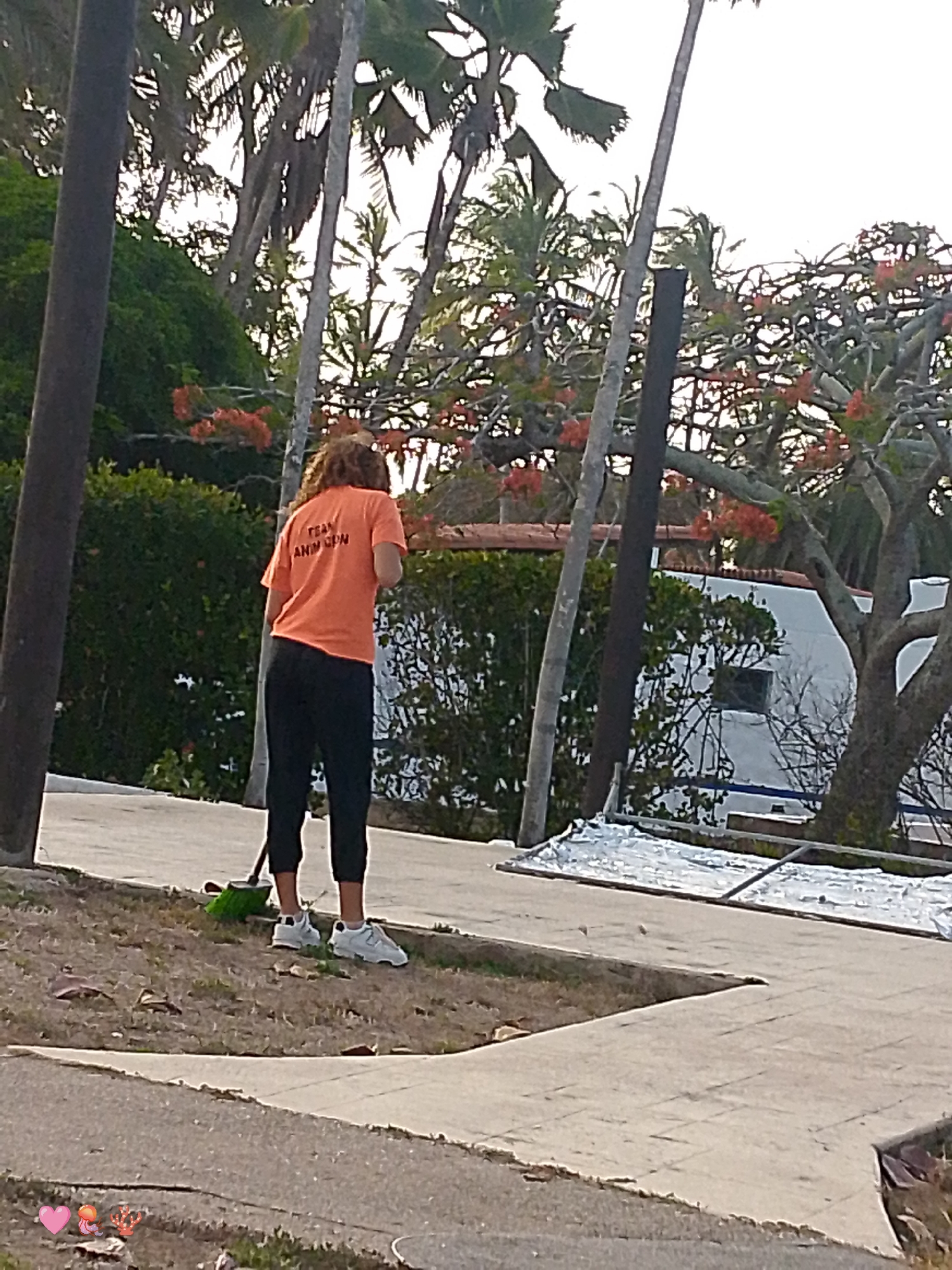 A woman wearing an orange T-shirt with the inscription "TEAM ANN CDN" and black pants is seen from the back, standing in front of a grassy patch next to a sidewalk. She is holding a green broom. In the background, there are trees and a white building with a blue trim. There is a tree with red flowers on the right side of the image. The photograph appears to be taken on a street in Playa El Agua, Venezuela.