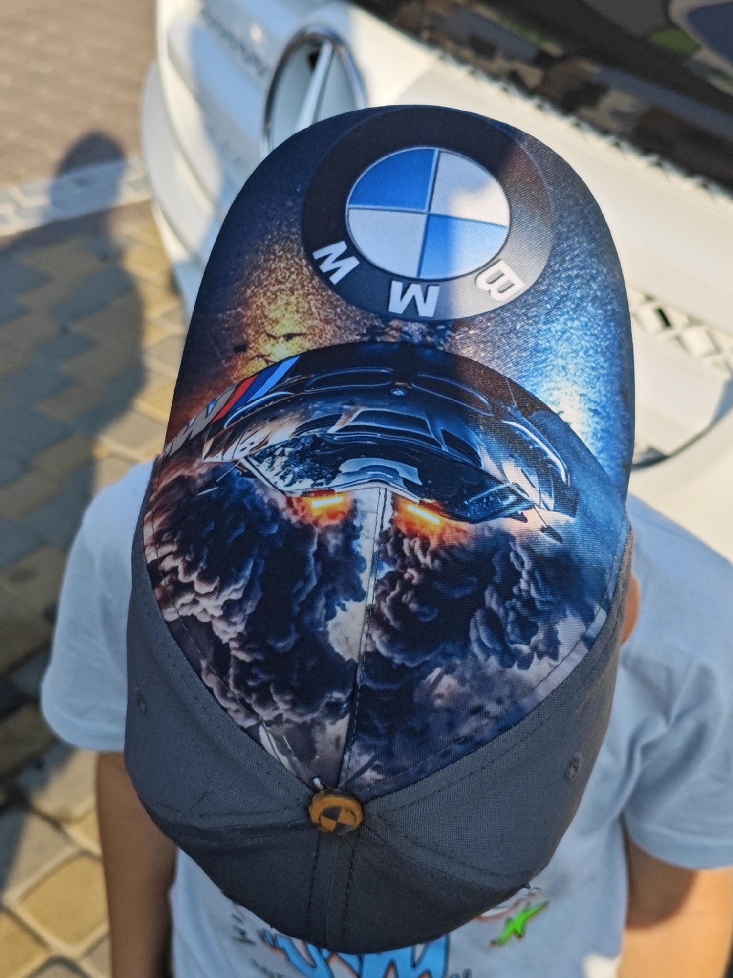 A high-angle shot shows a child wearing a gray baseball cap with a dynamic BMW M series car design. The cap's front panel is adorned with the BMW logo and a depiction of a sleek, blue M series car seemingly in motion amidst fiery orange exhaust flames and swirling smoke. The backdrop consists of a white vehicle, possibly a car, and a paved ground with a faint shadow of the child cast upon it. The setting appears to be outdoors, in a location that could be a parking lot or street in an urban environment, although the specific city and country are not identifiable from the image. The child's attire includes a light blue t-shirt.