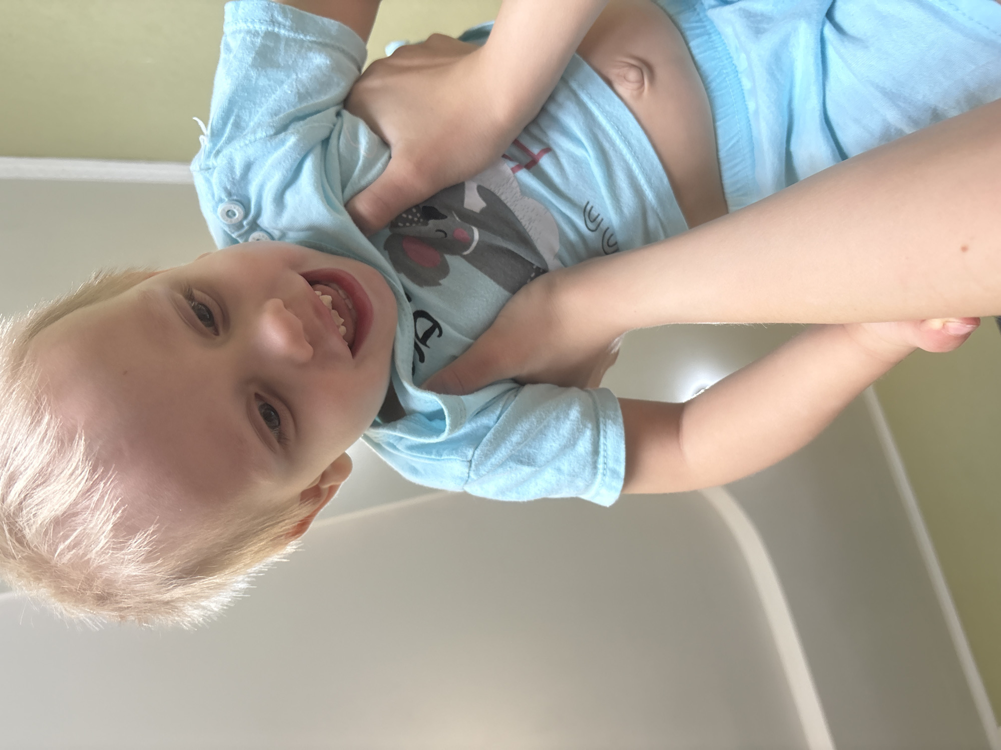 A young child with blonde hair is being held upside down, smiling, by an adult. The child is wearing a light blue shirt with a cartoon animal on it and light blue shorts. The adult's hands are grasping the child under the arms. The background consists of a white surface and a light green wall. The location is identified as Chaikovsky, Russia.