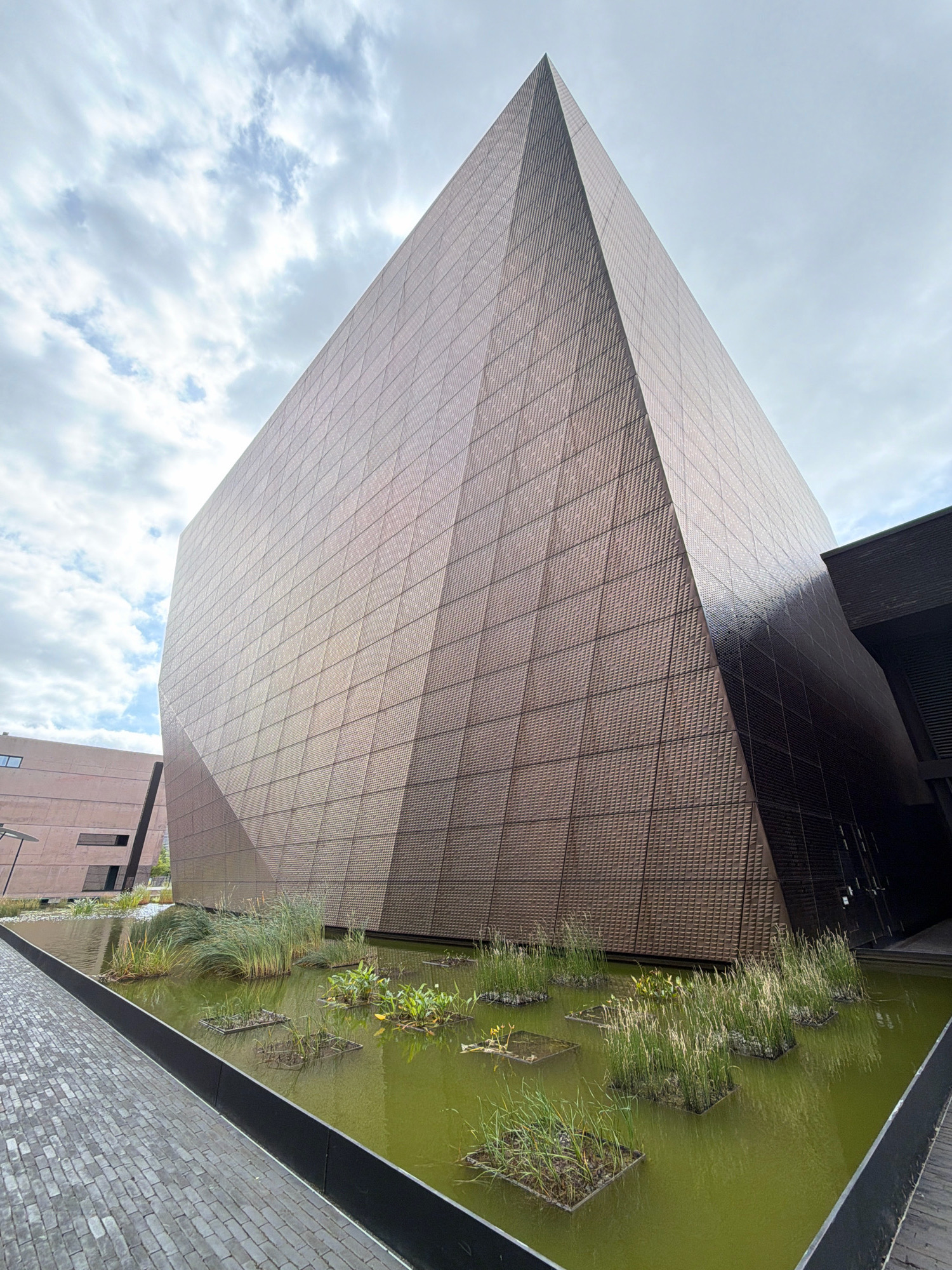 The image depicts a modern building with a distinctive geometric facade, characterized by a tiled surface that reflects the light. The building has a sharp, angular form, resembling a pyramid or a triangular prism, and is situated in an urban setting. In front of the building, there is a water feature with aquatic plants and grasses growing in shallow sections. The surrounding area includes a paved walkway with a brick-like pattern. In the background, another building with a more conventional rectangular design is visible. The sky is partly cloudy, with a mix of white clouds and blue sky. The scene is captured from a low-angle perspective, emphasizing the scale and architectural design of the main building. This building is the Maison de la Grande Region, which is located in Esch-sur-Alzette, Luxembourg.