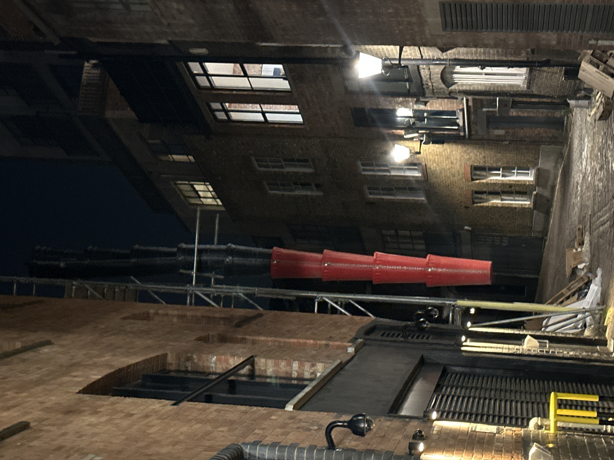 Captured from a low-angle perspective, the image reveals an alleyway in what appears to be London, United Kingdom, characterized by brick buildings and industrial infrastructure. Overhead, scaffolding and pipes are visible, including a series of red, cylindrical objects connected in a line, possibly serving as ducting or ventilation. The buildings flanking the alley feature windows, some illuminated, and an assortment of architectural details. Streetlights and lighting fixtures are affixed to the brickwork, and a metal grate is seen in the foreground. The environment is urban and industrial, potentially an old warehouse or manufacturing district undergoing renovation. No people are visible in the scene.