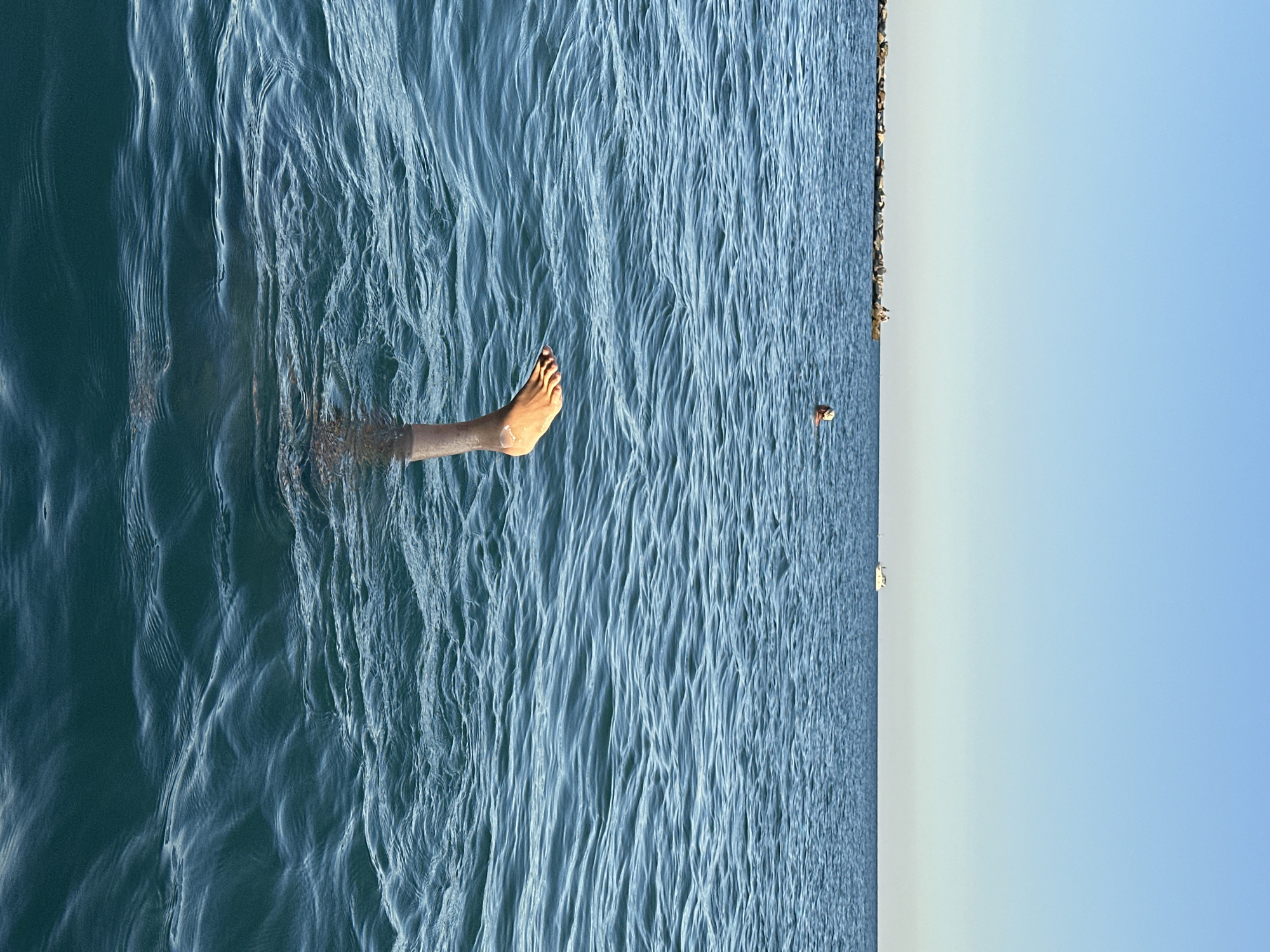 An aerial perspective shows a person's leg and foot submerged in the sea, with the toes pointing upward. The water is blue and rippled. In the distance, a jetty extends into the sea, with two people standing on it. Further out, a small boat is visible on the horizon. The sky is clear and blue. The scene is set in the Black Sea near Sochi, Russia.