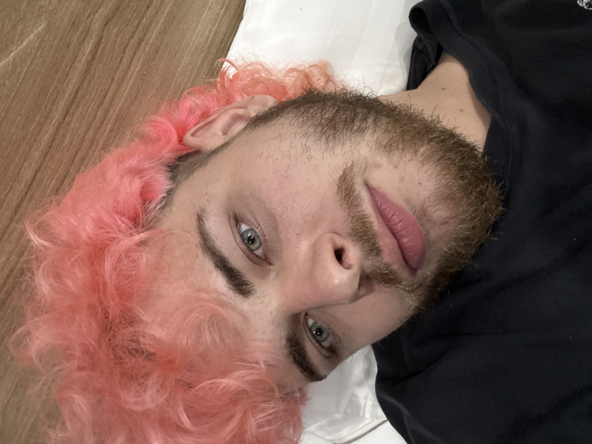 A close-up, diagonal portrait captures a male-presenting individual with striking, curly pink hair and a light brown beard/stubble. The person is lying down on a white pillow or sheet, with a wooden surface visible in the background, suggesting an indoor setting, possibly in Đà Nẵng, Vietnam, as per the provided context.
The subject has light blue-green eyes that gaze directly into the camera, conveying a neutral to slightly serious expression. Their lips are naturally full and slightly parted. The vibrant pink hair is tousled and frames the face, adding a playful contrast to the more earthy tones of their beard. Visible skin details include pores and minor blemishes. The individual is wearing a plain black t-shirt, which is partially visible on the right side of the frame. The lighting is soft and even, highlighting facial features without harsh shadows. There is no visible text in the image. The overall composition is a direct, intimate shot focusing on the person's unique characteristics.