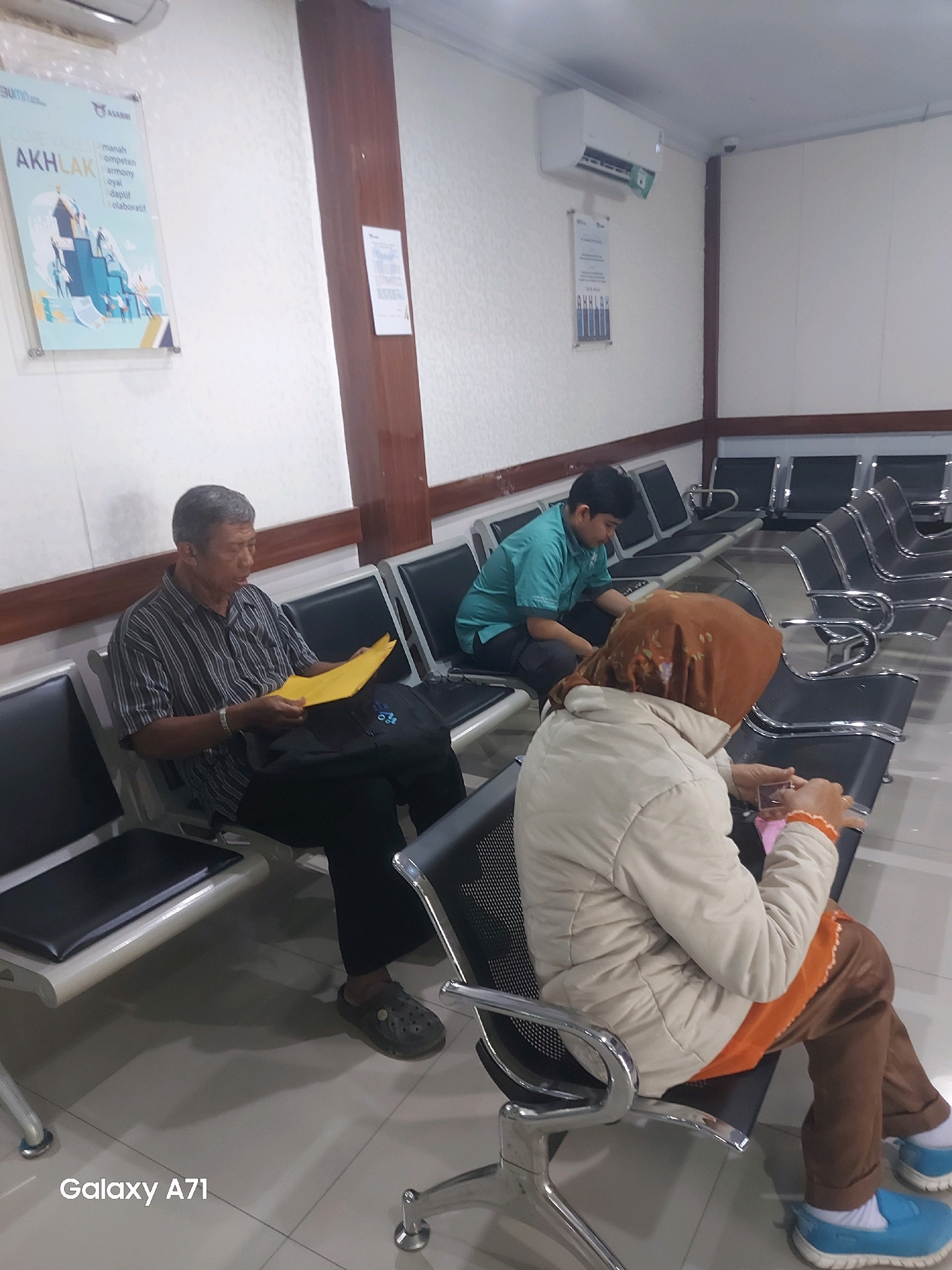 The image shows an indoor waiting area with several rows of empty and occupied seats. Three individuals are visible. In the foreground to the left, an older man with gray hair and a striped shirt is sitting and holding a yellow folder. To his right, a person wearing a light-colored jacket and a headscarf is seated, looking at their hands. In the middle ground, a young person in a teal shirt is sitting and looking down. The environment appears to be a government office or waiting room, with a poster on the wall that reads "AKHLAK" and "ASABRI," suggesting it could be a facility related to the Indonesian state-owned insurance company. An air conditioning unit is mounted on the wall. The floor is tiled. The image is watermarked "Galaxy A71" at the bottom left corner. The scene is likely in Makassar, Indonesia, given the presence of "ASABRI," a prominent Indonesian state-owned enterprise.