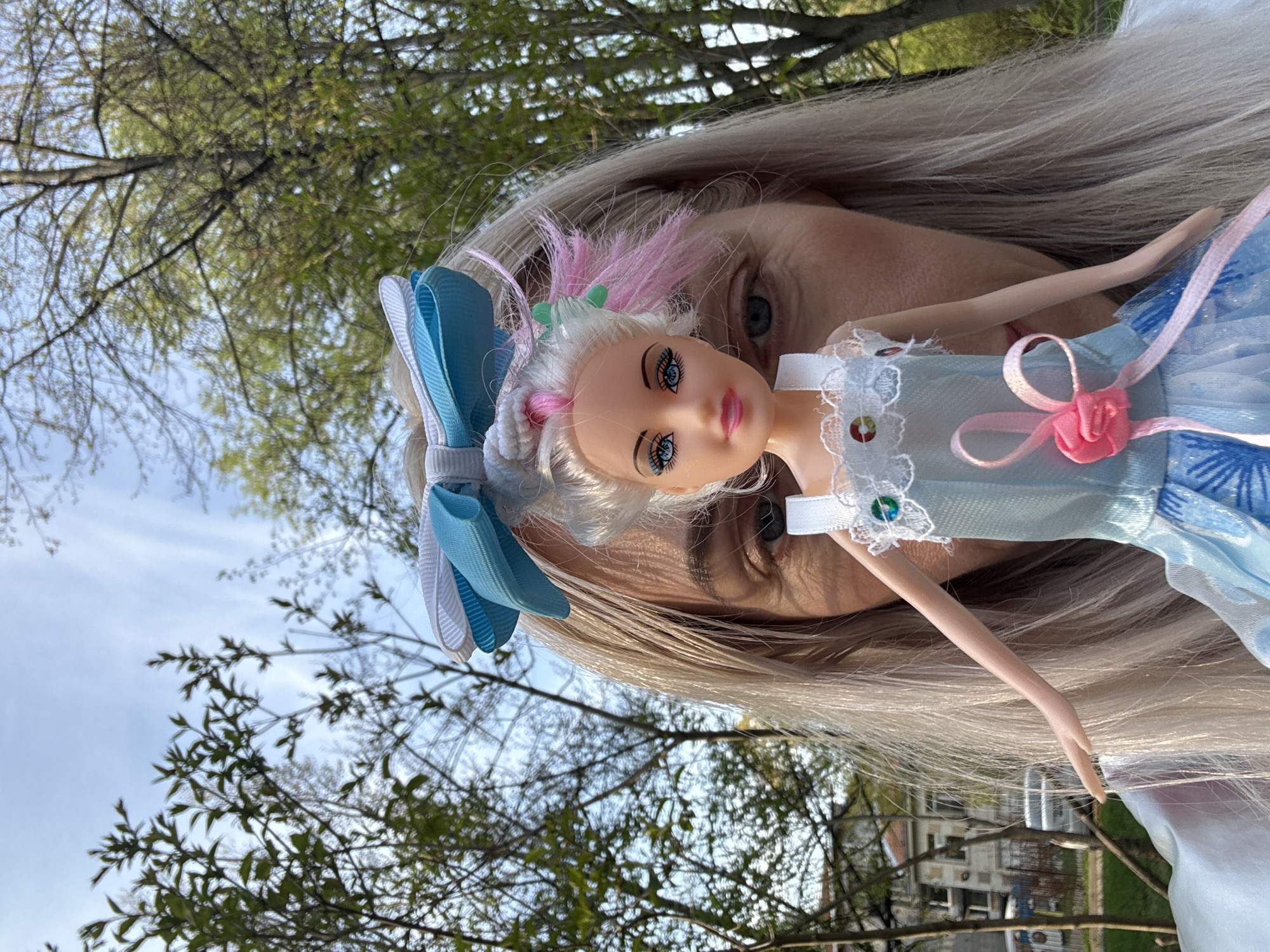 A woman with long blonde hair holds a fashion doll in front of her face, partially obscuring it. The doll has white hair with pink streaks, blue eyes, and wears a light blue dress with a pink ribbon detail. A blue and white ribbon adorns the doll's hair. The background features trees with budding green leaves against a bright sky. The location appears to be outdoors in a green space or park. The scene is captured from a low angle. There are houses in the background, although they are partially obscured. There is no evidence to confirm this scene is in the city of Saint Petersburg, Russia.