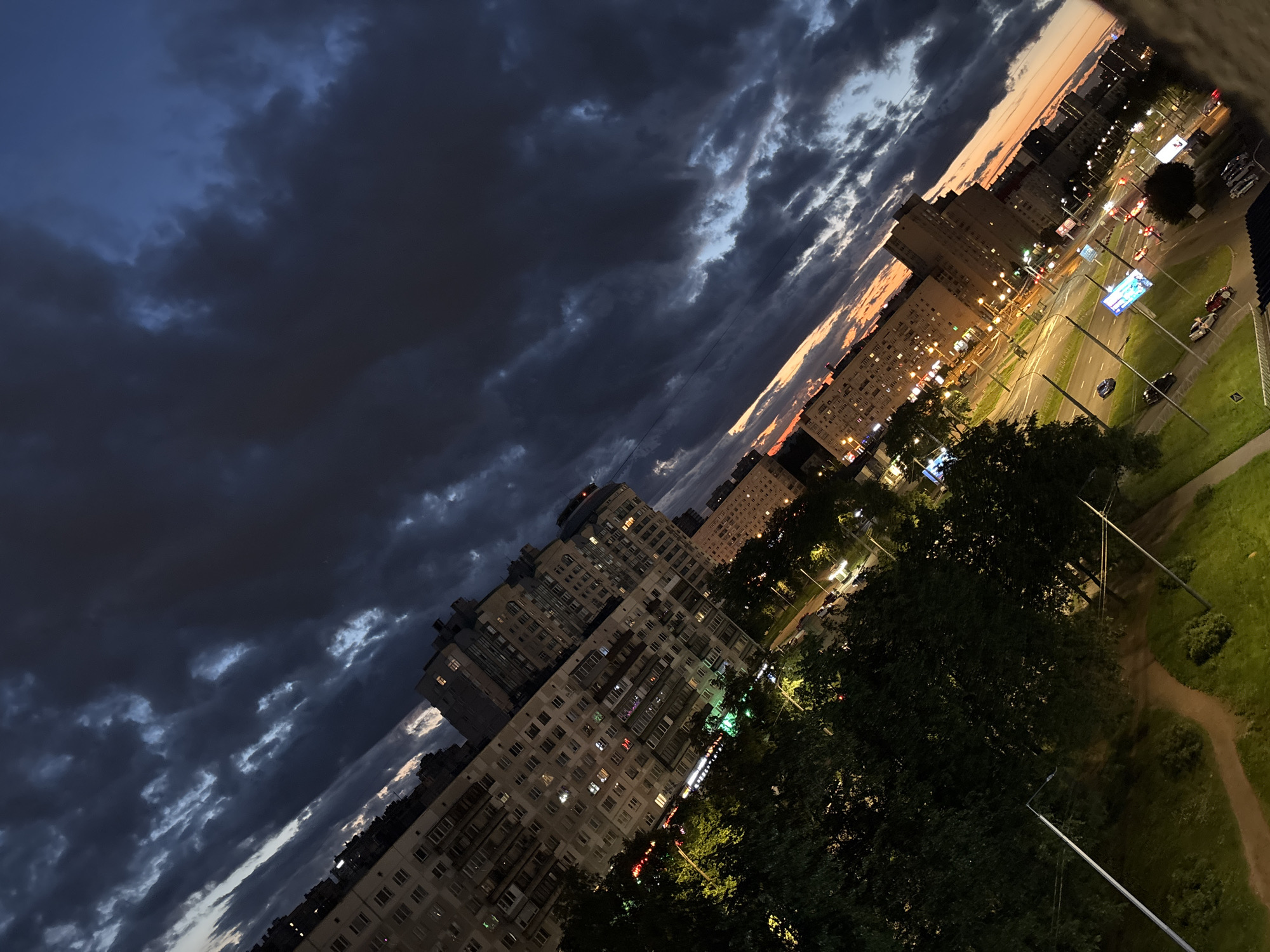 The image shows an urban landscape at dusk, viewed from an elevated perspective. The scene is dominated by a dramatic sky filled with dark, dramatic clouds, hinting at an approaching storm or the aftermath of one. Below the sky, a cluster of residential buildings with many windows, some illuminated, line the horizon. A street scene unfolds in the foreground, with cars visible on a road, suggesting a busy urban environment. Streetlights and illuminated billboards add to the nighttime ambiance. Lush green trees are scattered throughout the scene, their dark silhouettes contrasting with the artificial lights. The overall impression is one of a city at the cusp of night, with a moody and atmospheric sky.