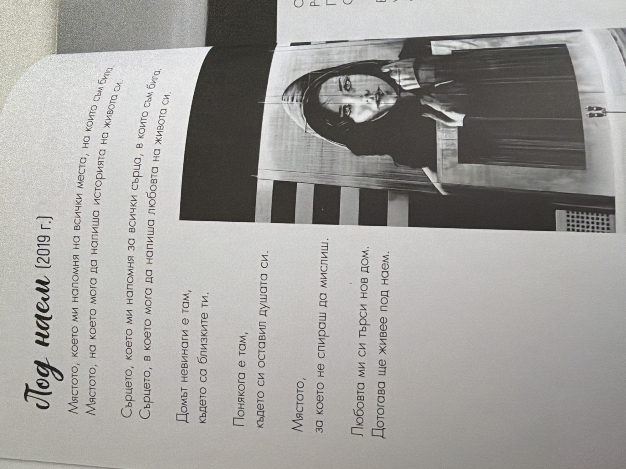 The image shows an open book with text on the left page and a black and white illustration on the right. The text on the left page is in Bulgarian and appears to be a poem. The title of the poem is "Под наем" (For rent), followed by "(2019 г.)". Below the title are several stanzas of text.
The illustration on the right page is of a woman's face, appearing to be a drawing or sketch. She is wearing a hooded garment and has a pensive expression. The background of the illustration is abstract, with vertical lines and shapes.
There is no information in the image that indicates a location in the city of Pazardzhik, Bulgaria. The image depicts a page from a book and an accompanying illustration.