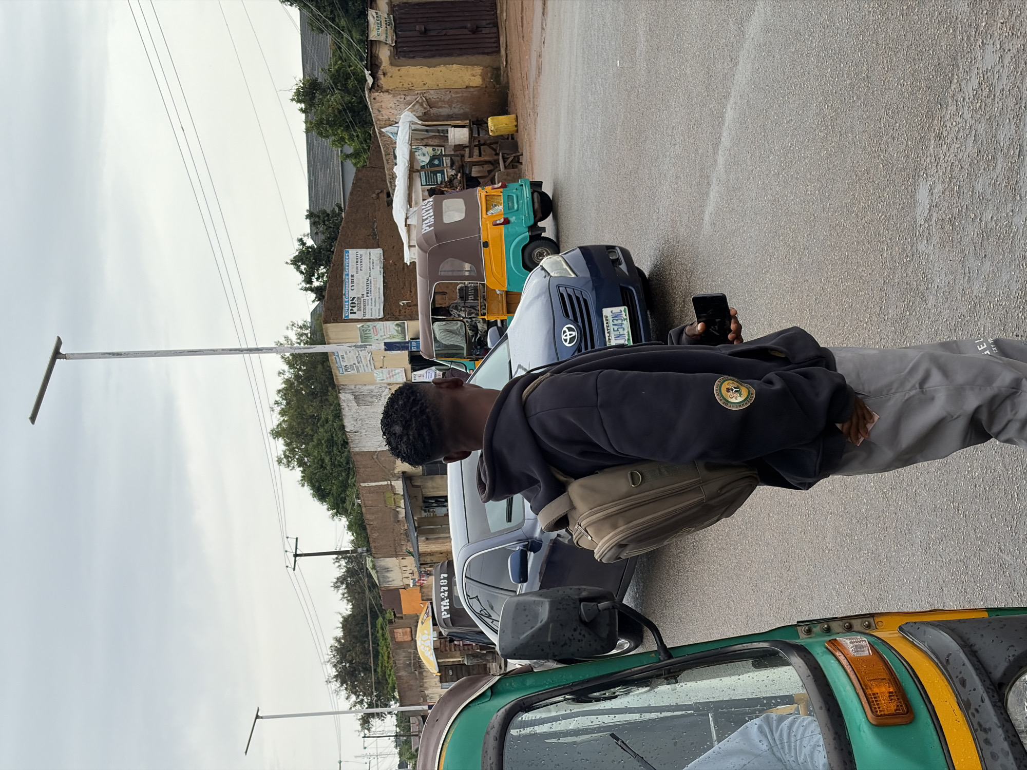 A dark-skinned male, seen from the back and right side, stands on a wet asphalt road in Jos, Nigeria, holding a dark smartphone in his right hand. He is wearing a dark hooded sweatshirt with an emblem on the left sleeve and light gray pants. A tan backpack is slung over his left shoulder. To his left, the front-left side of a green and yellow auto-rickshaw is visible, with a person's shoulder and arm inside. A dark blue sedan, with a license plate reading "JN-543NC", is positioned behind the auto-rickshaw and to the right of the young man, showing its front grille and headlights. Further in the background, a yellow and brown auto-rickshaw is parked on the right side of the road. The background features a street lined with buildings, some with storefronts and awnings. Power lines are visible overhead against a cloudy sky. The presence of puddles on the road indicates recent rain.