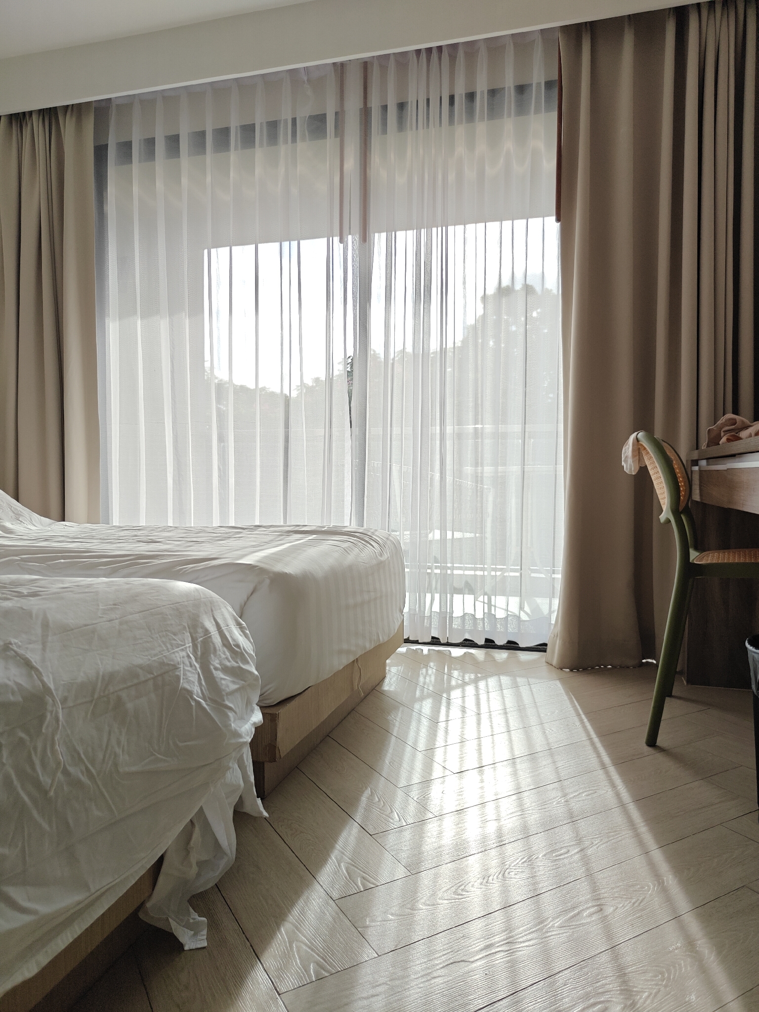The image shows a hotel room in Pattaya City, Thailand, with two beds and a large window covered by sheer white curtains and heavier beige drapes. Sunlight streams through the curtains, casting striped patterns on the light-colored wooden floor. To the right, a green cane-backed chair is positioned near a wooden desk. The beds are made with white bedding, and one bed appears to have its sheets slightly disheveled. The overall impression is of a bright, clean, and peaceful room.