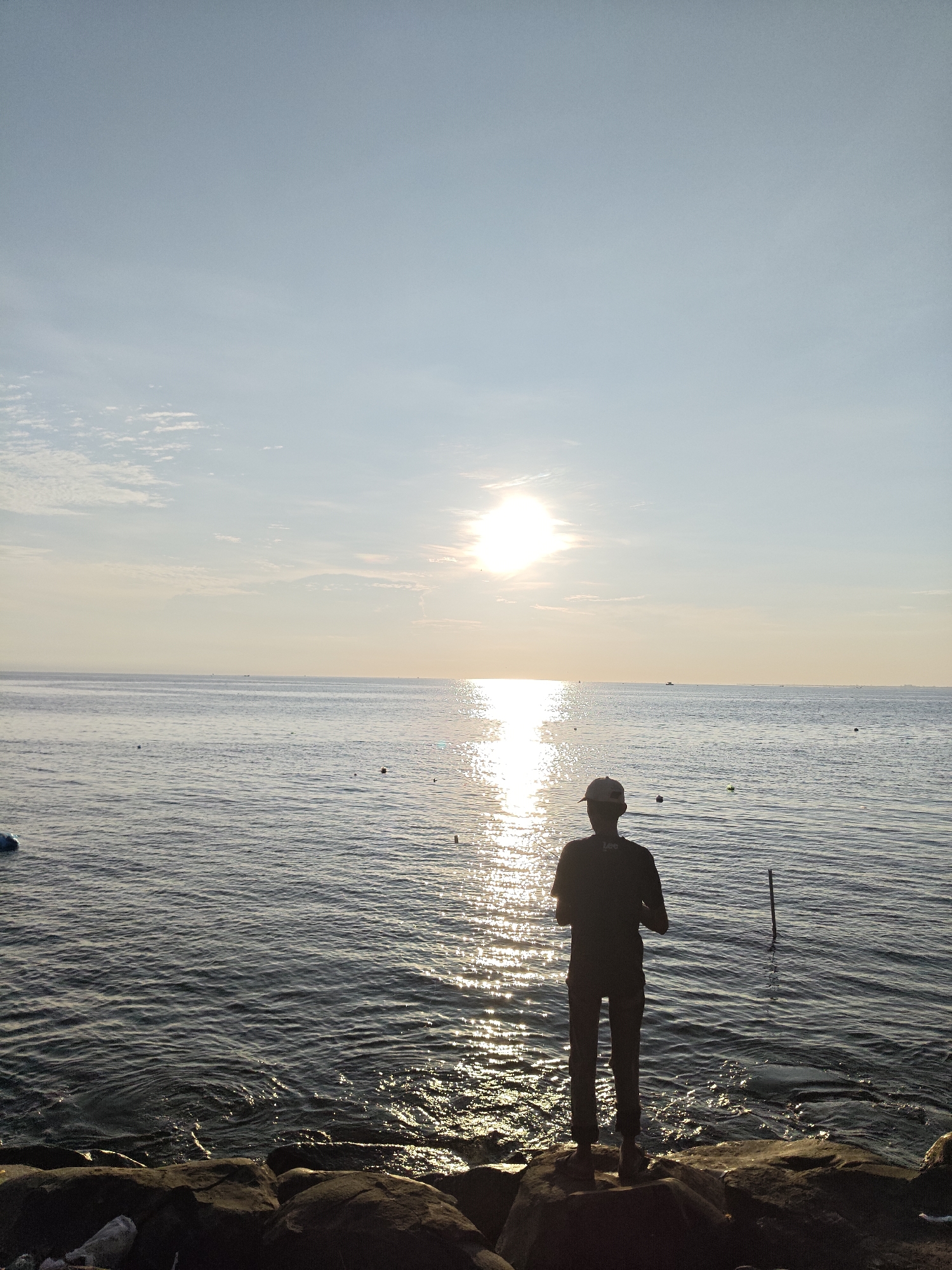 A person is standing on a rocky shore, looking out at a sea at sunset. The person is facing away from the viewer. The horizon is located at the center of the image. The image was likely taken in Lhokseumawe, Indonesia. The person is wearing a t-shirt, pants, and a cap. The sun is shining brightly in the sky, reflecting light off the water.