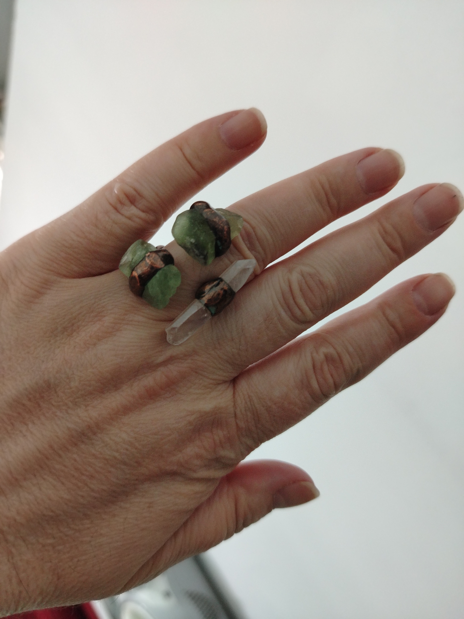 A close-up shot depicts a person's hand wearing three rings, each featuring a rough-cut stone secured with copper wire. Two of the rings feature green stones, while the third has a clear, pointed crystal. The background is plain and white. There is no information in the image to suggest the location is Al Mas'oudi, United Arab Emirates.