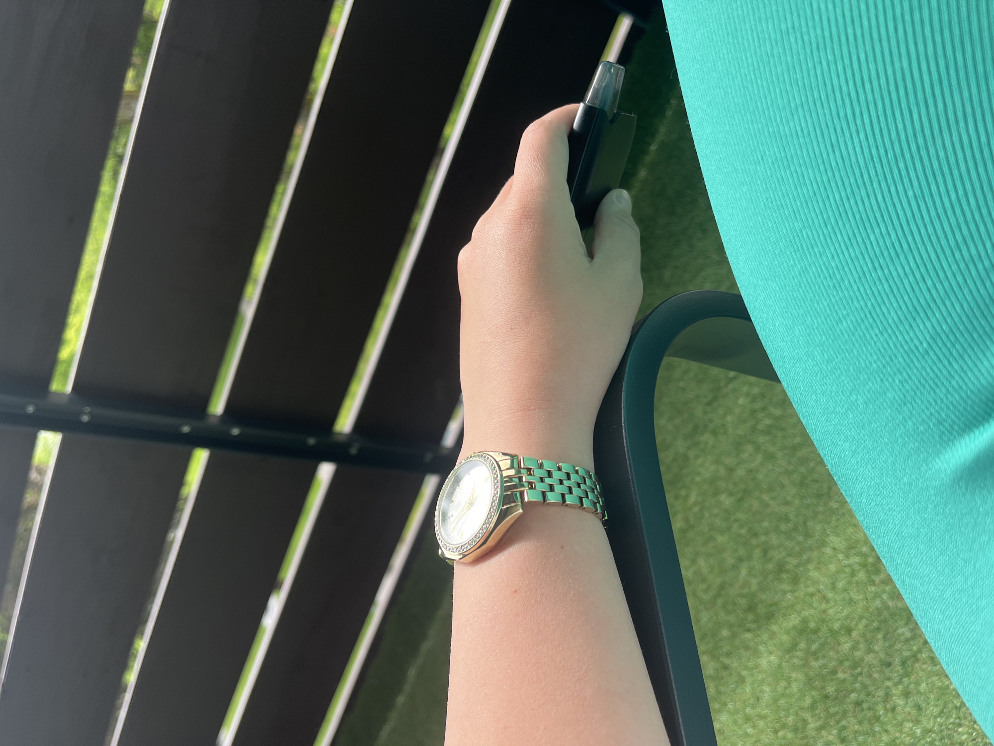 This image shows a close-up of a person's arm and hand. The person is wearing a gold watch with a crystal-embellished bezel and a teal-colored band. The person's hand is resting on a dark-colored object, possibly a chair. The background features a dark wooden fence with horizontal slats and a bright green, artificial turf-like surface, suggesting an outdoor setting. The overall lighting indicates it is daytime. There are no discernible landmarks or features that would allow for identification of a specific location within Garmisch-Partenkirchen, Germany.