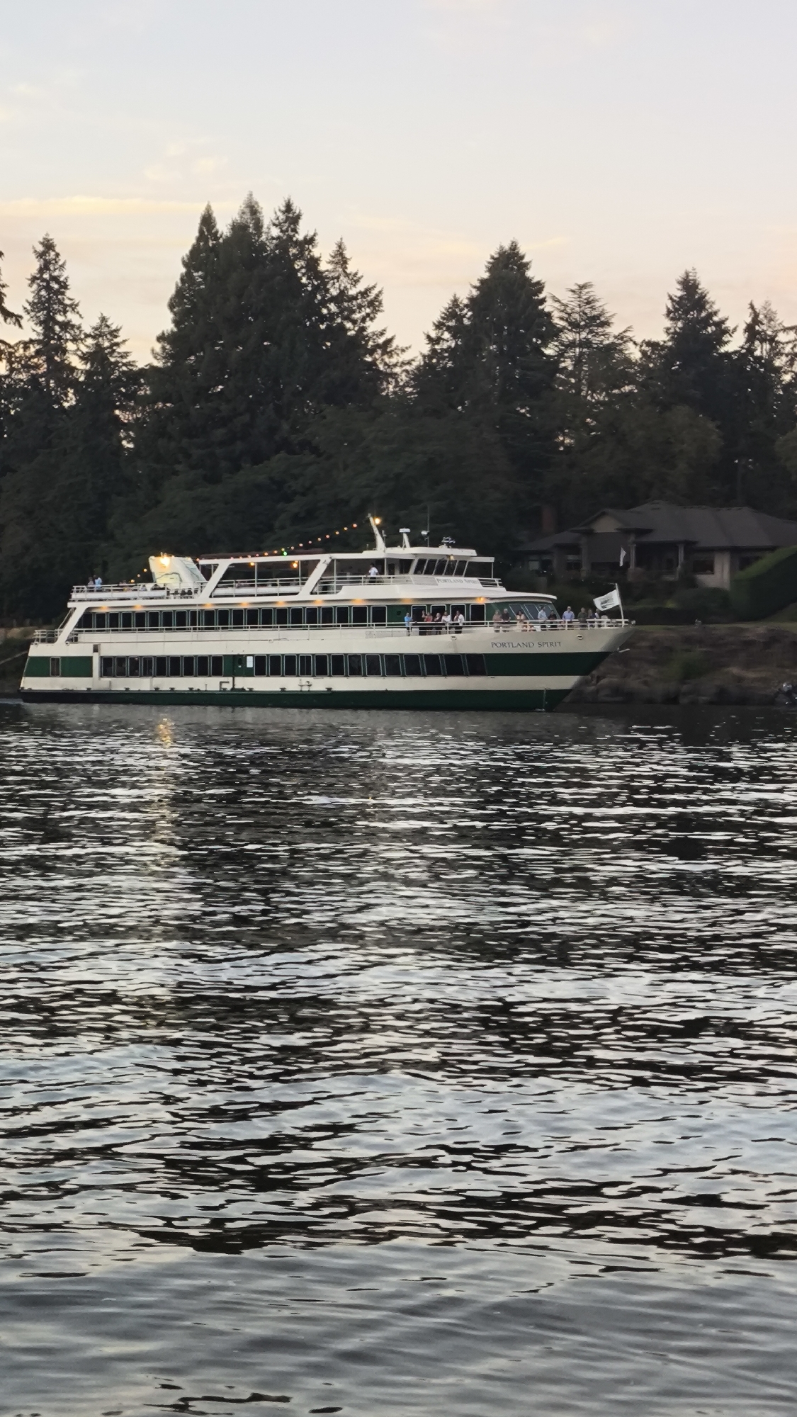 A large, white and green passenger boat named "PORTLAND SPIRIT" is docked in the foreground of the image. The boat is on a body of water that reflects the sky and surrounding trees. The water is choppy and shows ripples. In the background, there is a dense forest of tall, dark green evergreen trees. The sky above is a pale, hazy yellow and orange, indicating either sunrise or sunset. A large, modern-looking house with a dark roof is partially visible behind the trees on the right side of the image. Several people can be seen standing on the deck of the boat, and some lights are strung along the upper deck, suggesting it might be evening. The scene appears to be peaceful and serene, with the boat serving as the main focal point. The location is identified as Lake Oswego, United States of America.