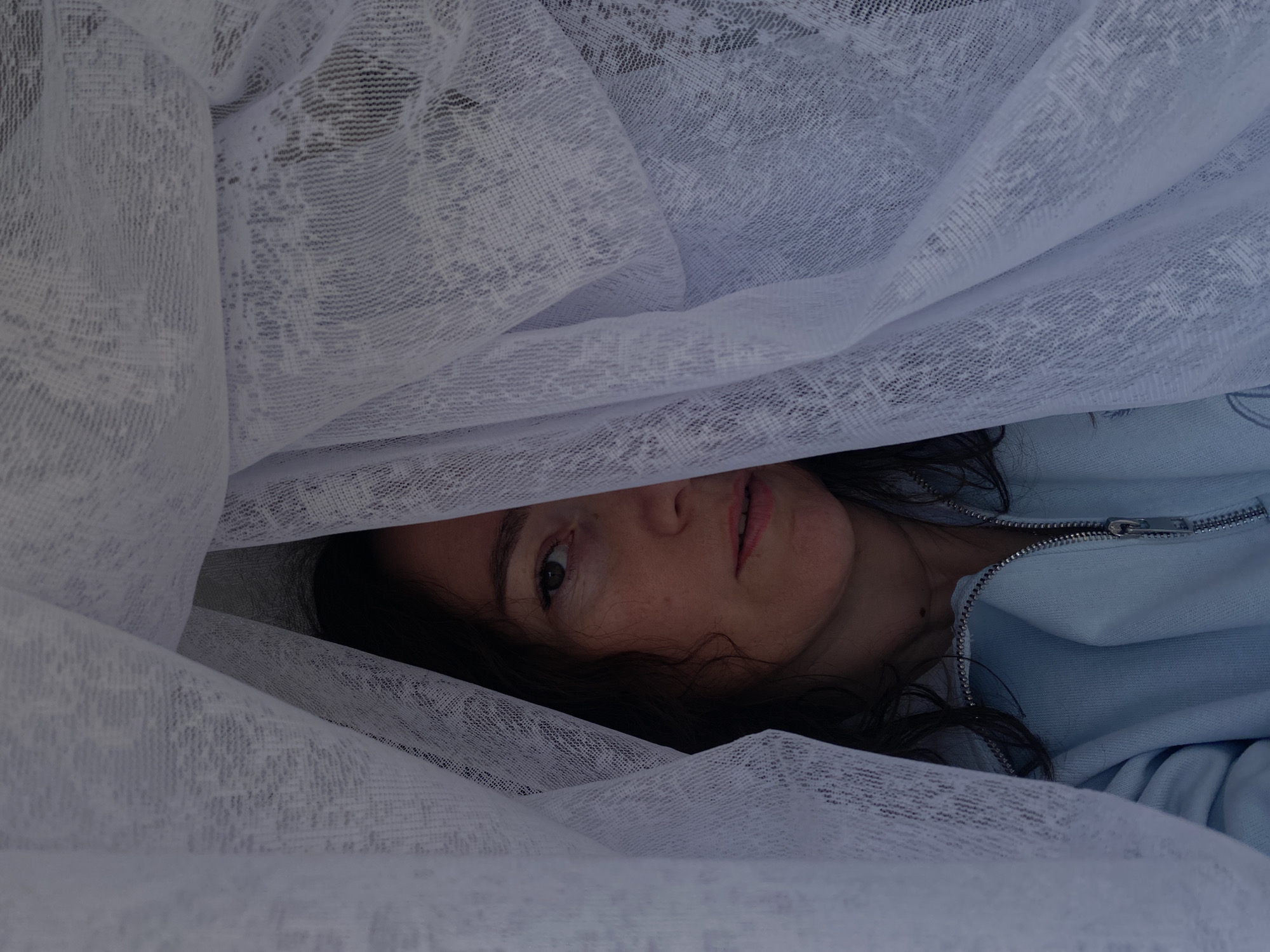 The image captures a close-up of a person's face, partially revealed from beneath folds of a light-colored, sheer fabric, likely lace or a delicate curtain. The person is positioned horizontally, with their left eye (from the viewer's perspective), nose, and mouth visible, looking directly at the camera. Dark, wavy hair frames the visible part of the face.
The translucent fabric, which covers most of the frame, is white or off-white and features a subtle, interwoven pattern that suggests lace or netting. It creates a soft, diffused lighting effect on the subject's face. Below the visible facial features and on the right side of the frame, a light blue garment, possibly a zippered jacket or sweatshirt, can be seen, with a silver-colored zipper running vertically.
The scene appears intimate and focused solely on the person and the interplay of light and shadow created by the fabric. There are no overt activities taking place beyond the direct gaze. The lighting is soft and even, suggesting an indoor setting or an overcast outdoor environment. No text is visible in the image. The location is provided as Ольховка, Russia, though no specific visual cues within the image confirm this.