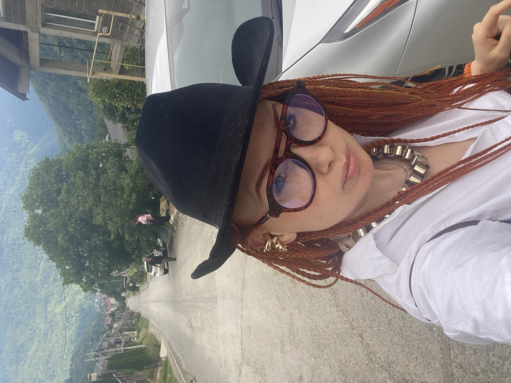 The media file depicts a close-up selfie of a woman with long, braided red hair and glasses. She is wearing a black hat and a white shirt with a chunky silver necklace. The background suggests an outdoor setting with a road, a car, and lush green mountains in the distance, characteristic of a location like Krasnaya Polyana, Russia. The lighting indicates it might be daytime, possibly overcast or with diffused sunlight. The woman's expression is relaxed. In the further background, there are some buildings and trees.