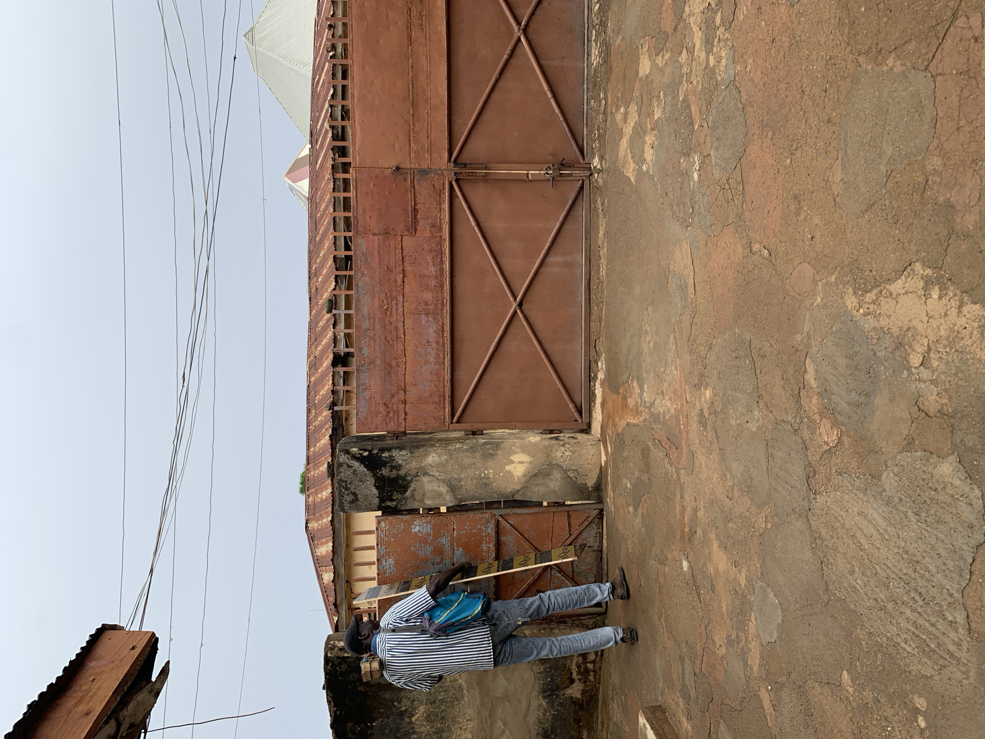 An eye-level, medium shot oriented on its side depicts a man in Rafin Par, Nigeria. He stands on a textured, cracked earthen surface next to a large, rusted metal gate in a dilapidated wall. The man wears a horizontally striped shirt, grey jeans, dark shoes, and a dark cap. A blue bag hangs from his shoulder, and he carries a stack of objects wrapped in white material. The gate is secured with a horizontal metal bar. Above the gate is a section of rusted metal with a ladder-like structure. Overhead, electrical wires cross a pale sky.