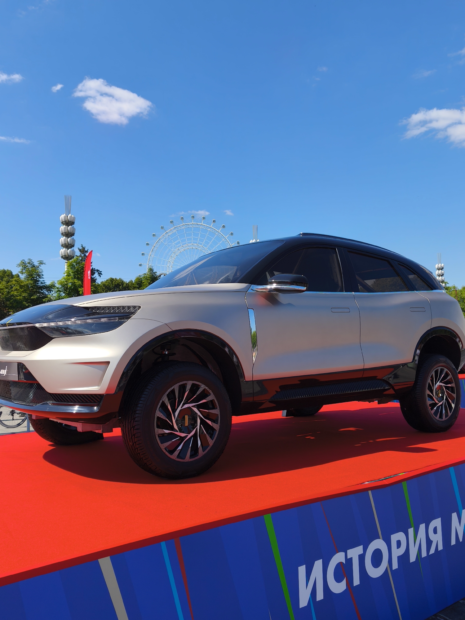 This is a photograph of a silver concept SUV parked on a bright red carpeted platform. The SUV has a black roof and black accents on the wheel arches and side skirts. The wheels are large with a distinctive silver and black spoke design. In the background, a Ferris wheel and a tall, modern sculpture are visible against a clear blue sky with a few scattered white clouds. Green trees line the horizon. A red flag is placed behind the front of the car on the left. The bottom edge of the image shows a blue banner with white Cyrillic lettering that appears to read "ИСТОРИЯ М" (History M). The overall scene suggests an outdoor exhibition or event, possibly in a public park or exhibition space in Moscow, Russia, given the presence of the Cyrillic text and the typical infrastructure of such venues. There are no people visible in the frame.