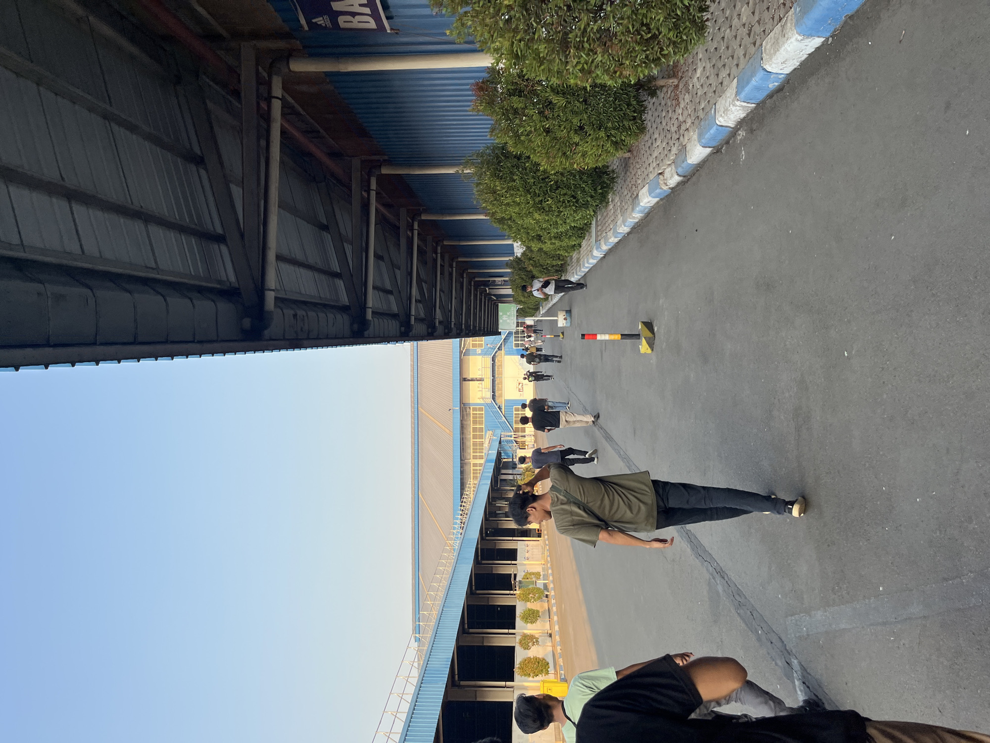 The image depicts an outdoor scene with a clear blue sky. In the foreground, a group of young men are walking away from the camera down a paved pathway. The pathway is lined with decorative bushes and a blue and white striped curb. On the right side of the pathway, there is a large building with a blue corrugated metal exterior and a metal roof. Part of a sign with the word "BA" is visible on the building. On the left side of the pathway, there is another building with a blue facade and a grey metal roof. The scene appears to be an industrial or commercial area. The location is identified as Pelang, Indonesia.