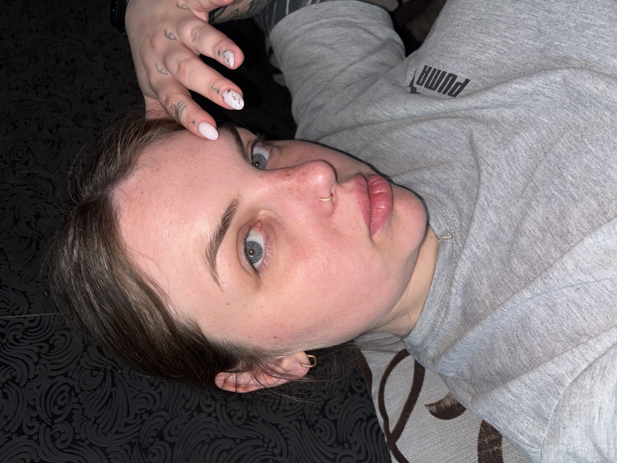 A young woman, likely in her late teens or early twenties, is depicted lying down in an indoor setting, possibly in Chornomorskoe, Ukraine. Her head is tilted, and her blue eyes gaze directly towards the camera with a neutral expression. She has light skin, noticeable freckles across her nose and cheeks, and dark hair framing her face.
Notable details include a small gold hoop piercing in her left nostril and a similar gold hoop earring in her right ear. Her right hand is placed on her forehead, revealing manicured nails; some are painted a light pale pink or white, while one features a white and grey marble-like design. Several small, black tattoos are visible on her fingers.
She is dressed in a light grey, long-sleeved top made of a soft, melange fabric. The brand name "PUMA" is clearly visible in black capital letters on the chest area of her top, appearing sideways and inverted from the viewer's perspective. The background consists of a dark, possibly black, patterned fabric, likely upholstery or bedding, featuring an intricate, swirling ornamental motif. The lighting is soft and even, suggesting an indoor environment. The image captures a casual, close-up portrait of the woman in a relaxed pose.