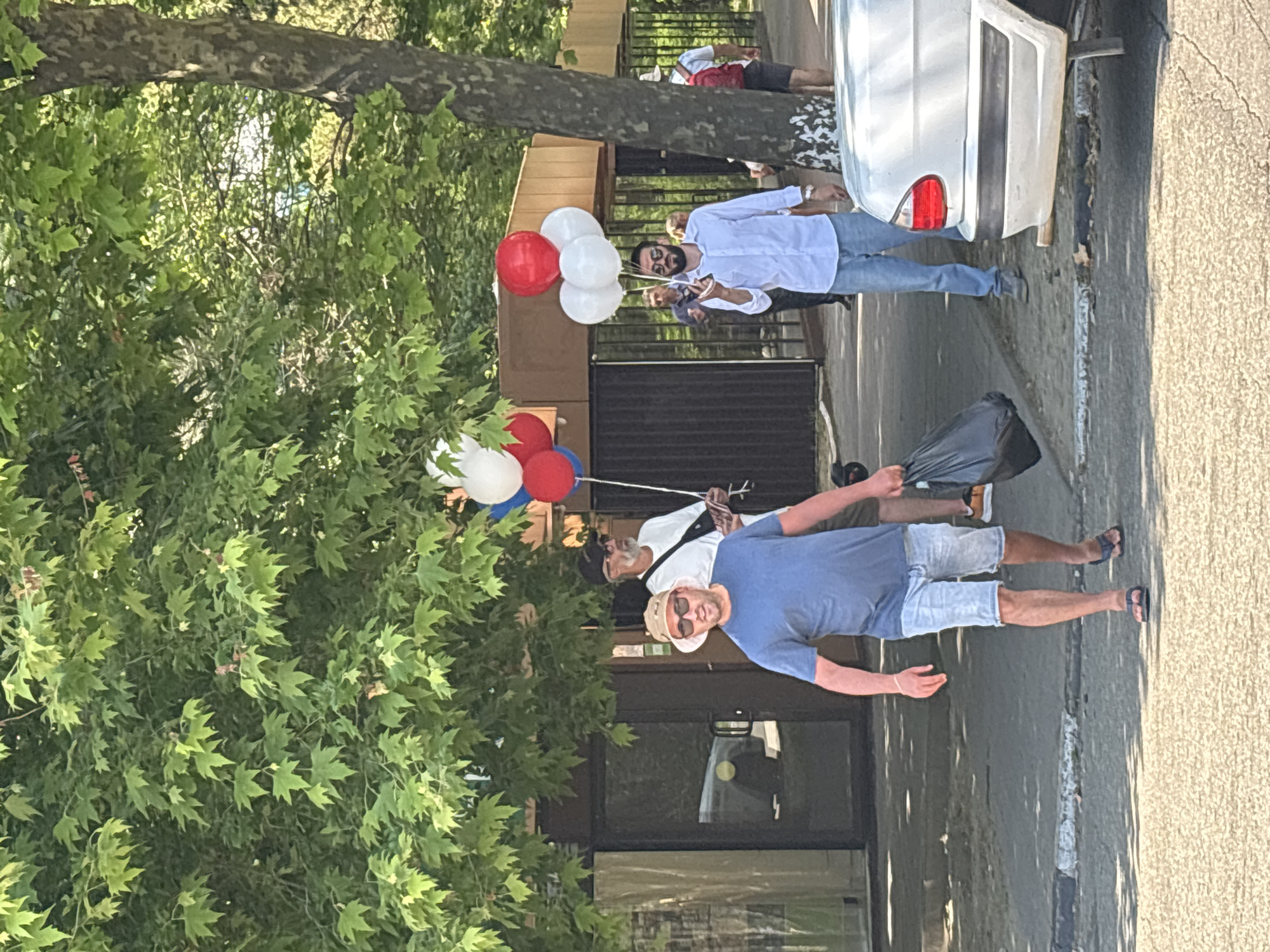Three men are walking on a sidewalk in front of a building with large glass doors and windows. A tree with lush green leaves shades the area. The man in the foreground wears a blue t-shirt, denim shorts, a straw hat, and sunglasses. He carries a black plastic bag in his right hand. Behind him, a man in a white t-shirt and a black baseball cap is visible. To the right of the foreground man, another man in a light blue collared shirt and blue jeans holds a bouquet of red, white, and blue balloons. A silver car is parked on the right side of the image, partially obscuring the view of other people and a fence in the background. The scene appears to be in Sochi, Russia, given the context of the image.
