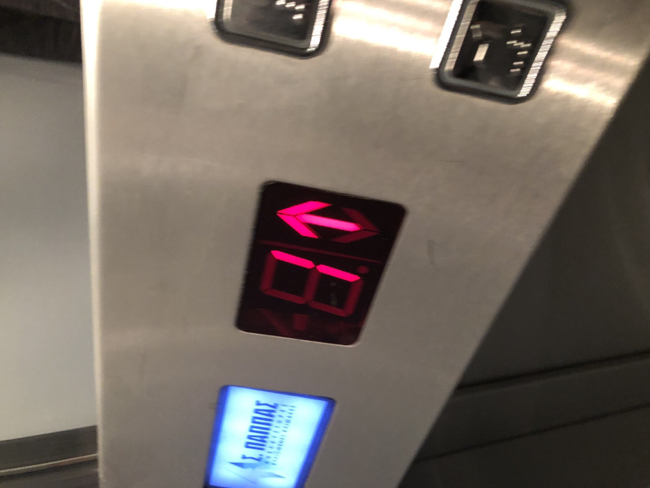 The image captures a close-up view of an elevator control panel, likely located inside an elevator car. The panel's surface is made of brushed metal. At the top, two square buttons are visible but unlit, with their symbols partially obscured.
The central and most prominent feature is a rectangular digital display illuminated in bright pink or red. This display shows a distinct left-pointing arrow at the top, and directly below it, the number "08". The arrow could signify the direction of the doors opening, a general directional instruction, or a specific status related to the elevator's current position or function on the 8th floor.
Below this main display is a smaller, rectangular display glowing with blue light. This blue display contains visible Greek text. The top line clearly reads "Σ. ΠΑΠΠΑΣ" (S. PAPPAS). Below this, smaller text states "ΑΝΕΛΚΥΣΤΗΡΕΣ" (Elevators) and "ΣΥΣΤΗΜΑΤΑ ΑΣΦΑΛΕΙΑΣ" (Security Systems). This text identifies the elevator manufacturer or service company as S. Pappas, confirming a Greek origin and aligning with the given location context of Moschato, Greece.
The scene is an indoor setting, with artificial lighting illuminating the panel. No individuals are visible, and the image solely focuses on the elevator's operational indicators and branding.