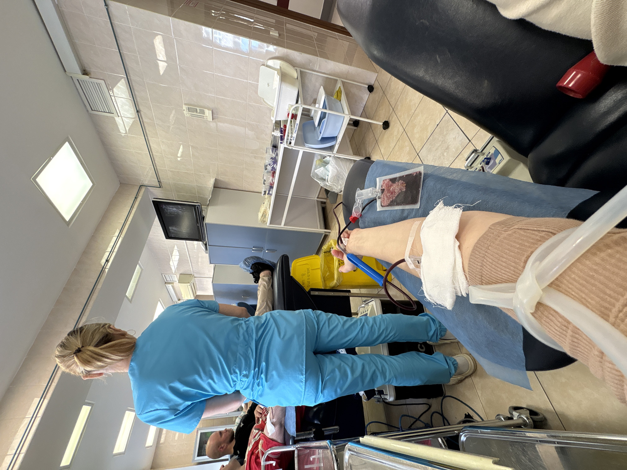 This image depicts an indoor scene within a medical facility, likely a blood donation or apheresis center in Saint Petersburg, Russia. The perspective is from a donor's viewpoint, looking across the room.
In the foreground, a person (the donor) is partially visible, resting in a black medical chair. Their right arm is extended, with a white bandage covering the venipuncture site. A clear tube runs from their arm to a collection bag, which contains collected blood or blood components. A light brown, possibly ribbed, sleeve covers the donor's upper arm, and a blue medical drape is spread over their lap.
In the midground, a medical professional wearing light blue scrubs stands, facing away from the camera, attending to the equipment. They have blonde hair tied back. Nearby, a white metal utility cart with shelves holds various medical supplies, including bottles, containers, and a blue basin. Faint Cyrillic text, possibly "РГМ ПРЕПАР" or "РКМ ПРЕПАР" (likely indicating "preparations"), is visible on an item on the cart, confirming the Russian context. A yellow medical waste bin is also present on the floor. In the background, another person wearing a red shirt is seated in a similar chair, suggesting they are also a donor or patient.
The setting is characterized by light-colored tiled walls and floors, indicative of a clean, sterile medical environment. The ceiling features several square and rectangular fluorescent light panels, along with air vents. A dark-screened monitor or television is mounted on a wall to the left. The overall scene suggests a routine medical procedure, likely blood or plasma donation, conducted in a professional and functional clinical space. The time of day is not discernible, as the room is lit by artificial light.