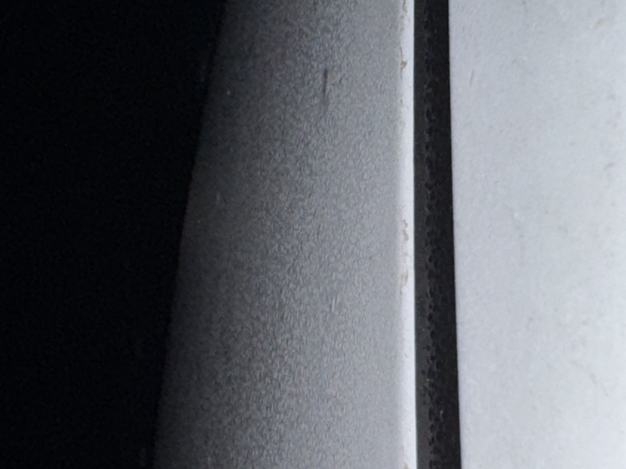 The image is a close-up, abstract view of textured surfaces. On the left, a deep black area dominates, suggesting a shadow or a dark material. Transitioning to the right, there's a textured gray surface with a fine, granular appearance. This gray section has subtle variations in tone and appears to have very faint vertical marks or imperfections. Further to the right, a sharp, vertical line separates the gray surface from a lighter, off-white area. This white surface also exhibits a subtle texture, though it is less pronounced than the gray. The lighting appears to be diffused, creating soft transitions between the different areas. There is no visible text. The scene is devoid of any identifiable objects, people, or a clear setting, making it difficult to infer an activity or specific location beyond the abstract textural qualities presented. The monochromatic palette and focus on surface detail contribute to an overall minimalist and perhaps industrial aesthetic.