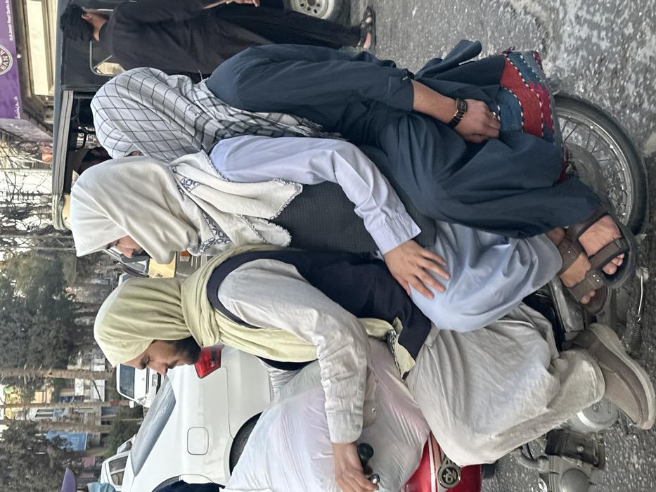 In Quetta City Tehsil, Pakistan, three men are depicted riding a red motorcycle on an urban street. The scene is observed from a slightly elevated, side-profile perspective, with the motorcycle traveling leftward relative to the frame.
The driver, positioned at the front of the motorcycle, has a dark beard and wears a light beige head covering, a light-colored long-sleeved tunic (kurta), a dark vest, and light-colored trousers (shalwar). He is wearing brown slip-on shoes. His left hand is visible near the handlebar, and a large, white, partially translucent bag is positioned in front of him on the motorcycle.
The middle passenger sits directly behind the driver. He wears an off-white head covering with a patterned border and scalloped edge, a light blue long-sleeved tunic, a dark vest, and light blue trousers. He wears dark brown sandals, and his right hand rests on the driver's left thigh.
The rear passenger is seated behind the middle passenger. He wears a white and grey checkered head covering, a dark blue long-sleeved tunic, and dark blue trousers. He also wears dark brown sandals (partially visible) and a black watch on his left wrist. The rear seat of the motorcycle is covered by a patterned textile featuring red, blue, and white colors.
The street surface is asphalt, exhibiting some wet or stained patches. In the background, other vehicles are visible, including a white sedan and a black auto-rickshaw. Storefronts line the street, one displaying a purple sign with text that includes "BANK" and "M.A. Jinnah Road Quetta." Bare-branched trees and utility poles with overhead wires are also present in the background. The lighting suggests daytime conditions.