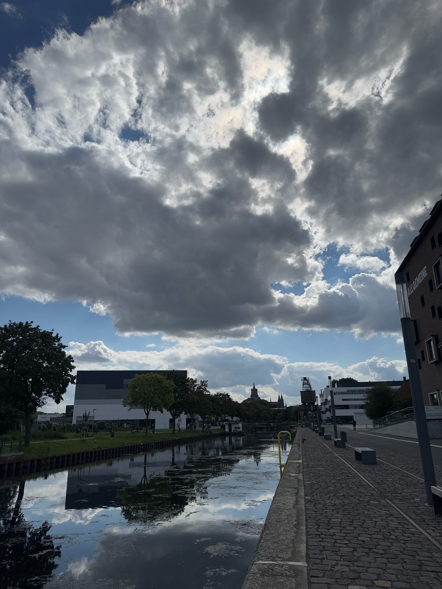 The image depicts a canal-side scene in Cleves, Germany. A paved walkway lines the right side of the canal, with several dark benches and what appear to be old train tracks embedded in the cobblestones. On the far right, a brick building with windows is visible, featuring signage that reads "ALLGEMEINE". Across the canal, to the left, is a modern, multi-story building with a dark facade and a row of trees in front. Further in the distance, various other buildings, including what seem to be church spires and a large industrial crane, are visible. The canal water reflects the sky and surrounding structures. The sky is dramatic, filled with a mix of dark and light clouds, with patches of blue sky showing through. The overall atmosphere is overcast but bright. There are no visible people in the immediate foreground or engaging in any actions.