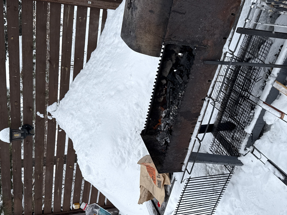 This image depicts an outdoor winter scene in Sysert, Russia. The foreground features a dark, rusted metal charcoal grill (brazier) resting on snow-covered ground. The grill contains partially burned charcoal with visible embers and small flames. To the left of the grill, on a snow-covered surface, a crumpled brown paper bag with red text and a QR code is visible. A clear plastic bottle containing a blue liquid, capped in white, is partially seen on the far left, near a snowdrift. The grill structure includes lower metal support racks and several metal skewers on the right side, all partially covered in snow. In the background, a substantial pile of white snow is mounded against a dark brown wooden fence constructed of horizontal slats. A wall-mounted lantern, partially obscured by snow, is affixed to the fence. Bare tree branches are discernible behind the fence. No individuals are present in the scene.