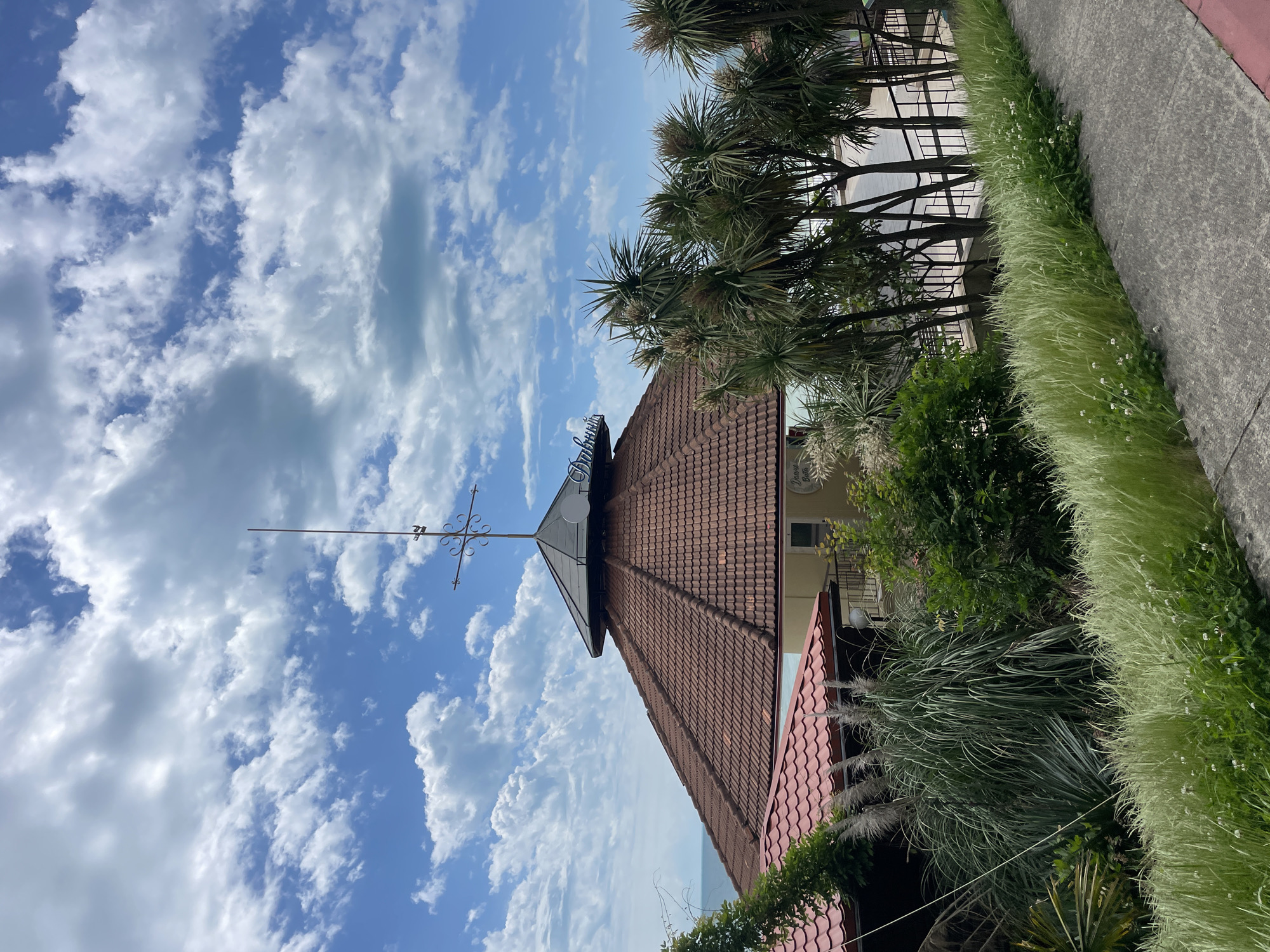 The image shows a building in Sochi, Russia. The building has a red tile roof with a spire topped with a cross. The sky is blue with white clouds. There are trees and greenery in the foreground. A sidewalk runs along the edge of the image with tall grass growing along it. The building appears to be situated near the sea, based on a glimpse of water visible in the background.