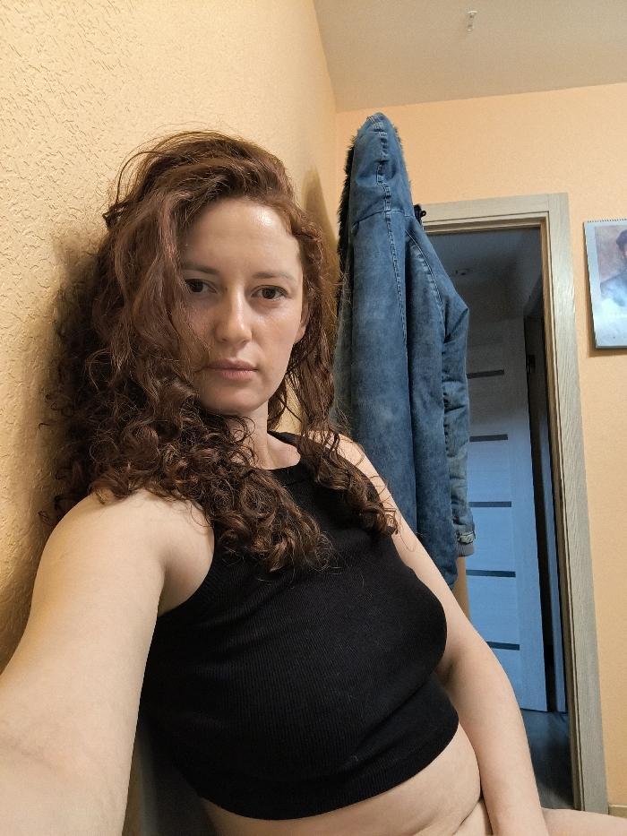 This image captures a medium-shot selfie of an adult female within what appears to be a domestic interior in Saint Petersburg, Russia. The woman, who has long, curly, reddish-brown hair, looks directly at the camera with a neutral expression. She is wearing a black, ribbed tank top that reveals her midriff. Her left arm and shoulder are pressed against a textured, light orange wall, while her right arm extends out of frame, presumably holding the camera. To the right of the woman, a blue denim-style jacket with a visible fur-lined hood hangs on a hook, partially obscuring a light-colored wooden door frame. The door beyond is slightly ajar, revealing a darker interior space with horizontal striped features. Further to the right on the wall, a small framed picture or calendar depicting a bust portrait is visible. The lighting is consistent with indoor artificial illumination.