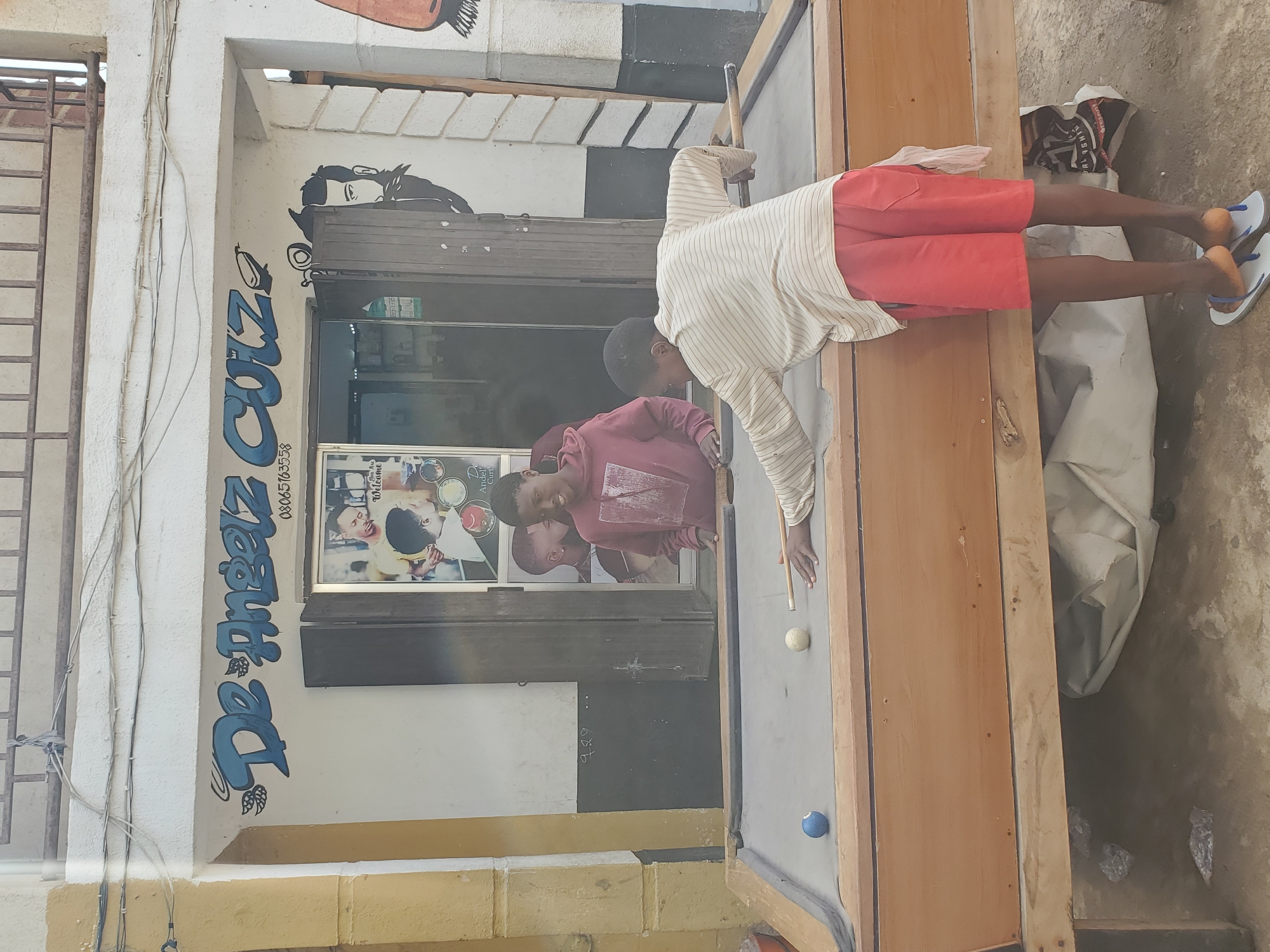 In a sun-drenched outdoor setting in Jos, Nigeria, two young boys are engrossed in a game of pool. The scene unfolds in front of a building with a white facade adorned with blue graffiti that reads "De Princez Cut" and a phone number. A framed picture on the wall displays portraits of men, possibly representing the services offered. One boy, wearing a striped long-sleeved shirt and red shorts, is positioned behind the pool table, cue in hand, ready to take a shot. The other boy, in a maroon long-sleeved shirt, watches intently, a slight smile on his face. The pool table, showing signs of wear, is set on a gray surface with a few billiard balls visible, including a white cue ball and a blue ball. Beyond the table, discarded items and debris suggest a less-than-pristine environment. The bright daylight indicates it is daytime, and the overall atmosphere seems relaxed and playful.