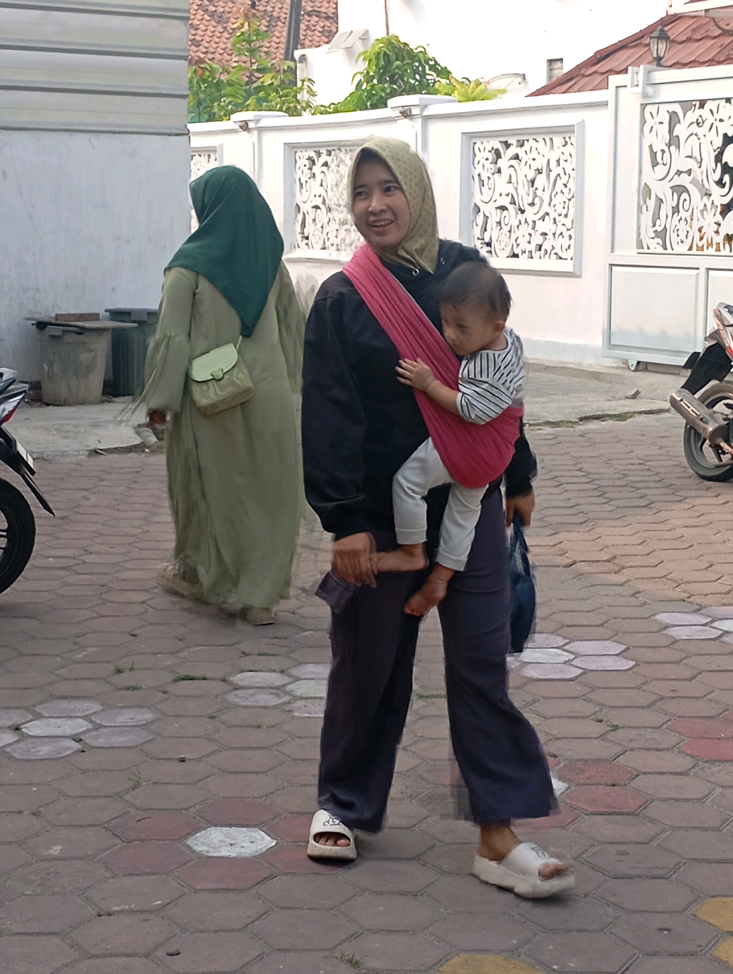 In the city of Babelan, Indonesia, a woman wearing a light green hijab and a long flowing green dress walks away from the camera. She carries a light green shoulder bag. In front of her, a woman wearing a yellow-green hijab and a black jacket is carrying a baby in a pink sling. The baby is wearing a striped shirt and light pants. The woman is wearing dark pants and white platform sandals. She is holding a dark blue bag in her left hand. The ground is paved with hexagonal bricks. In the background, there is a white building with decorative white panels. A motorcycle is parked on the right side of the image.